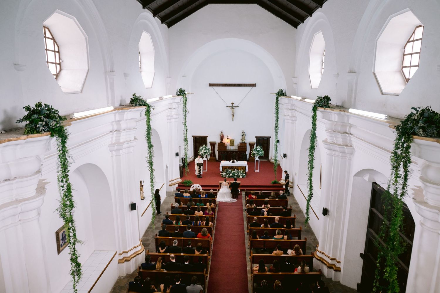 Interior view of classic white church with red carpet aisle and decorated with greenery for wedding ceremony.