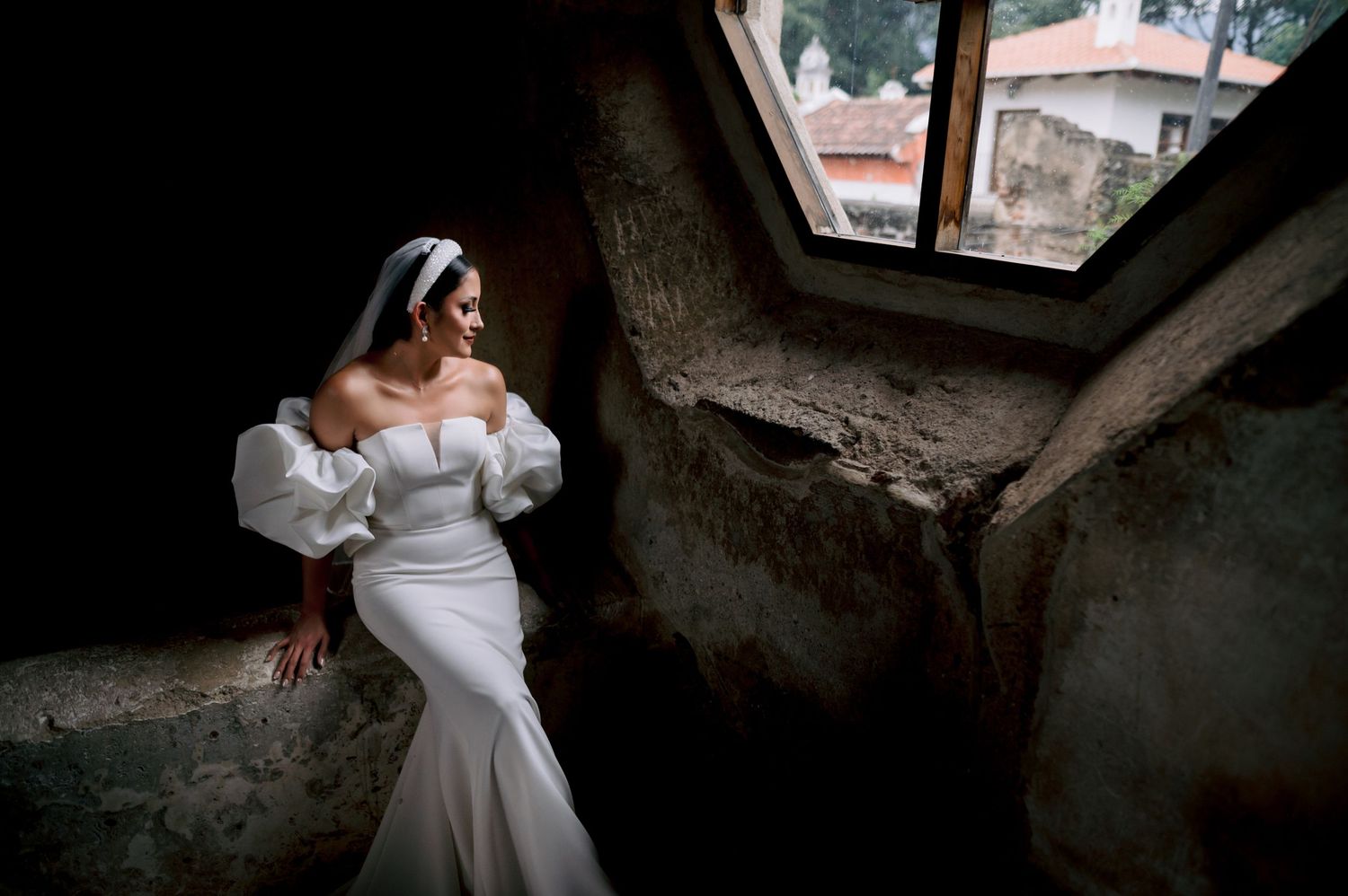 Bride in elegant white dress poses by a window in a dramatic architectural setting.