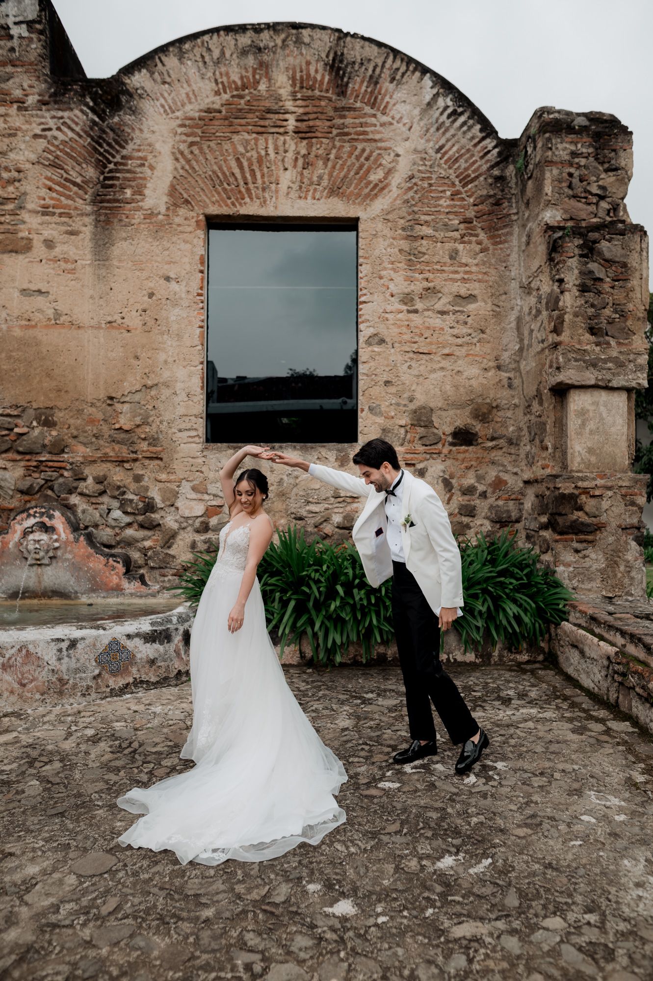 A newlywed couple playfully poses against ancient stone walls with a modern window backdrop.