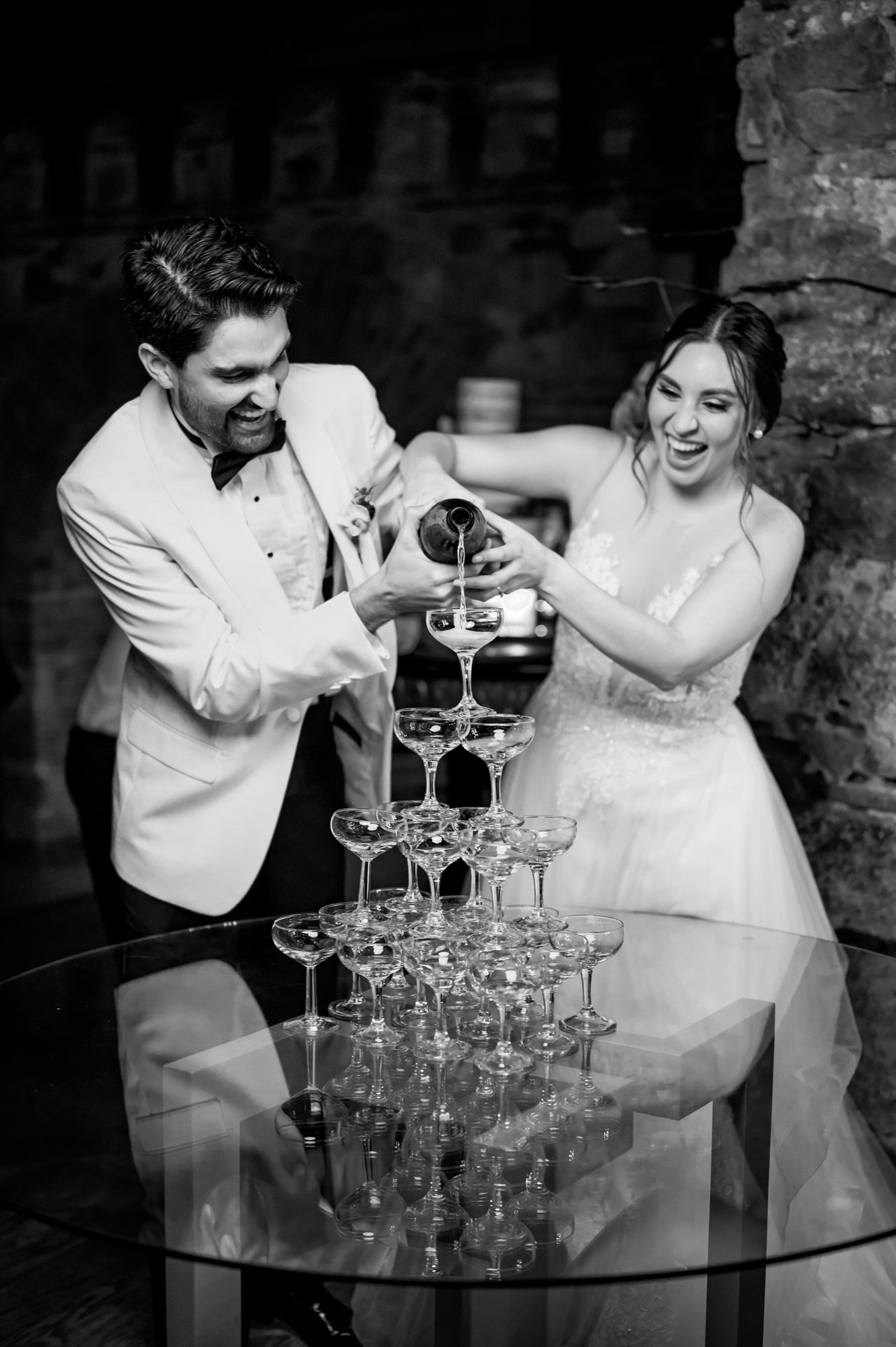 Black and white photo of couple pouring champagne into tower of glasses at wedding reception.
