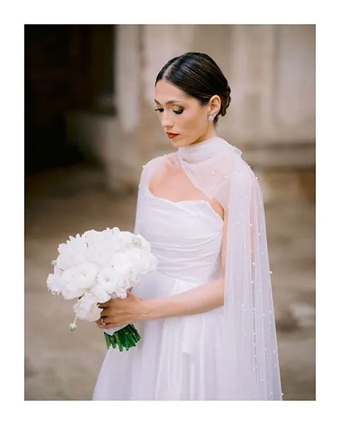 An elegant bride in a white high-neck gown with sheer cape detail poses with a bouquet of white peonies.