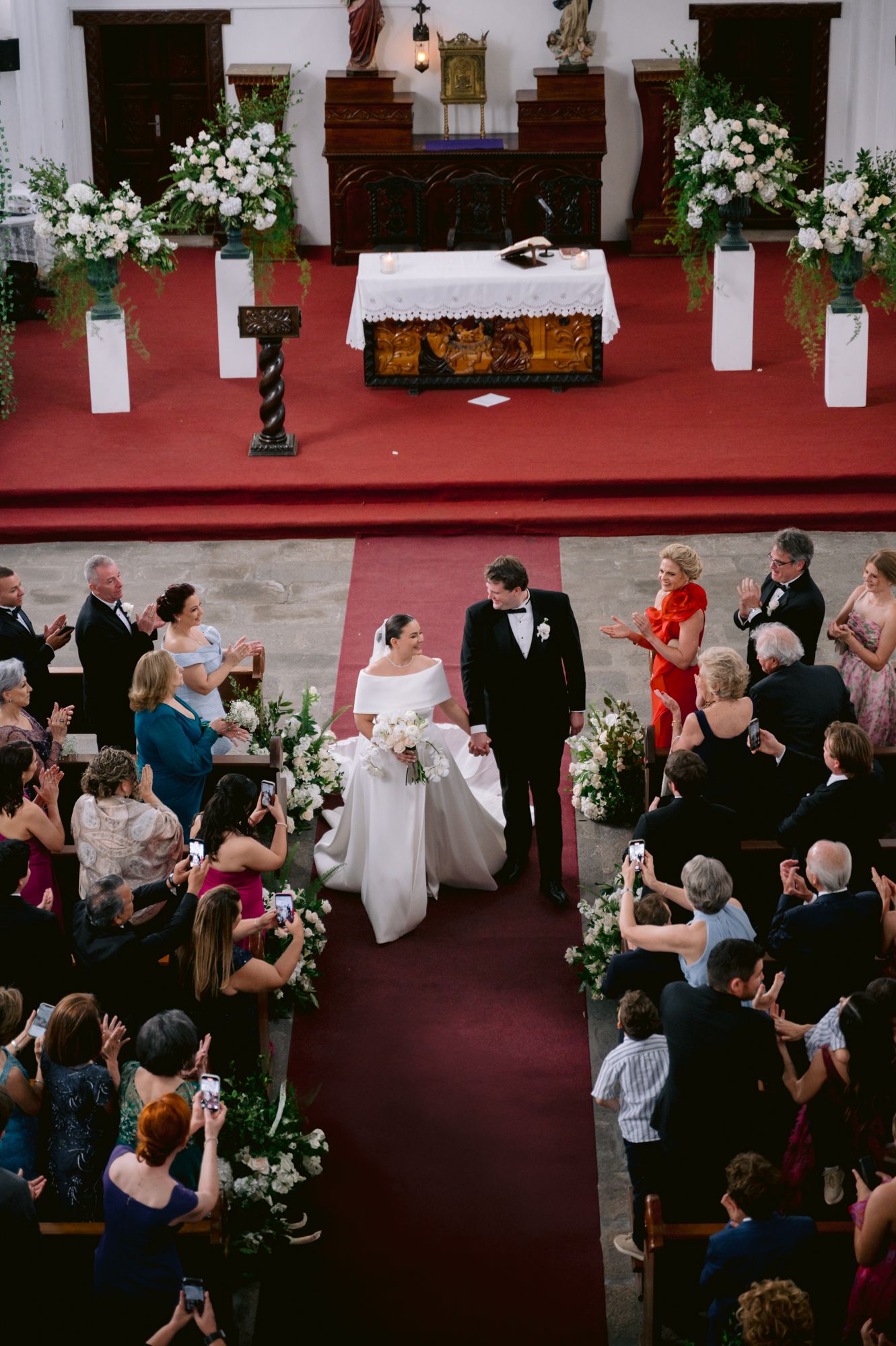 Aerial view of a church wedding ceremony with guests watching the couple amid white floral decorations.