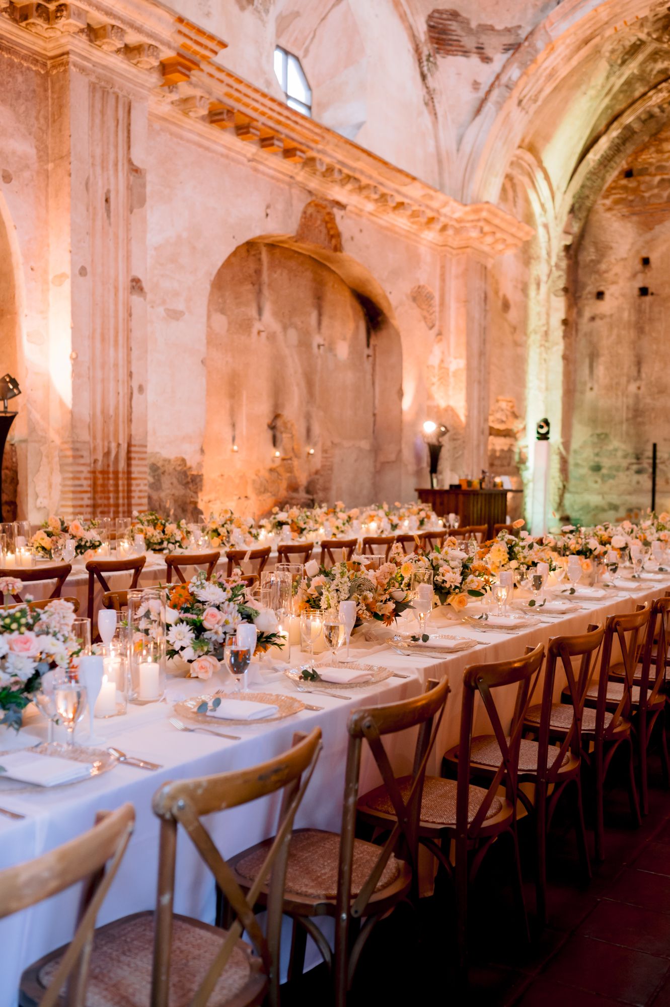 Romantic reception dinner setting with wooden chairs and floral centerpieces in historic chapel.