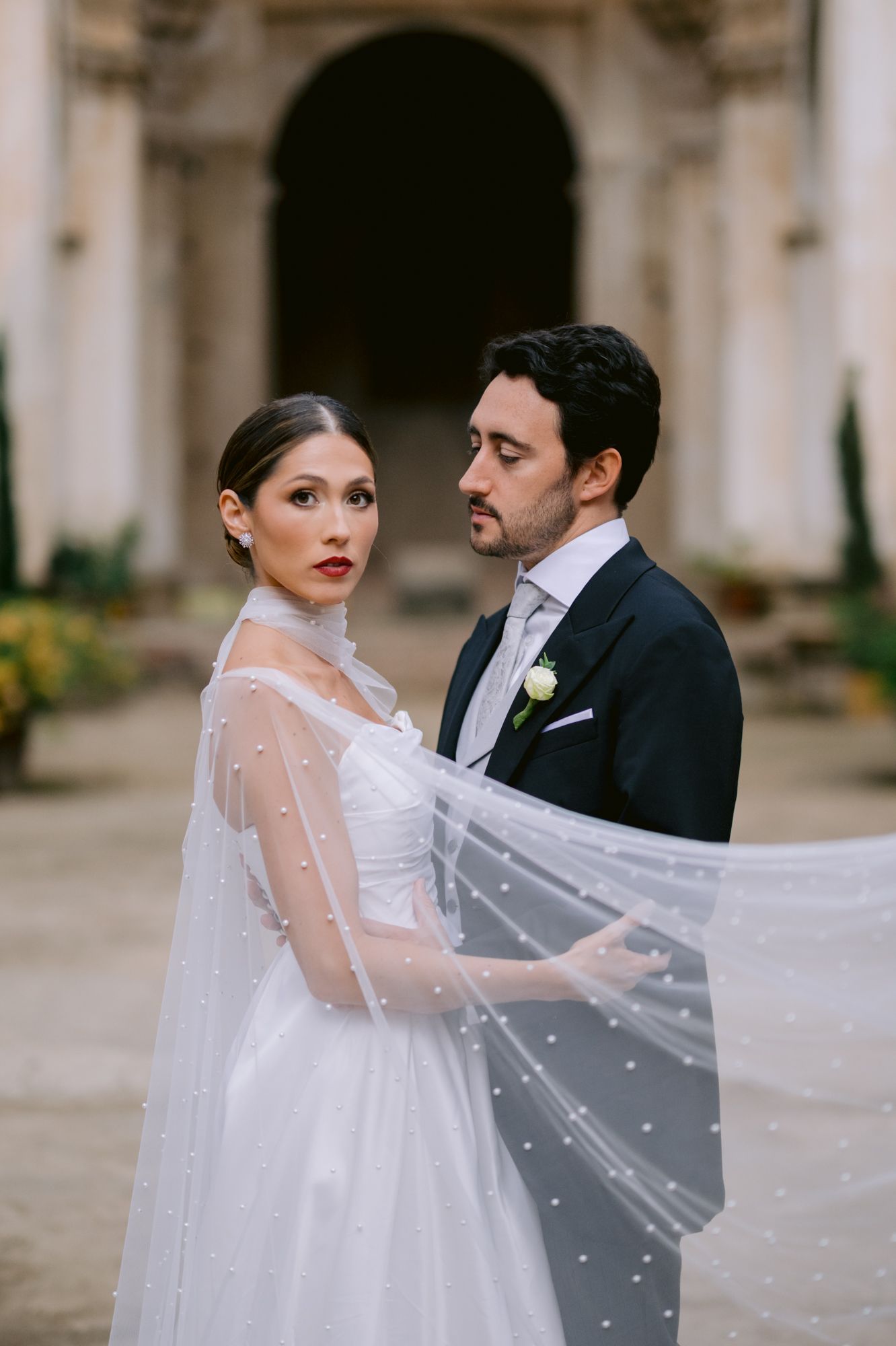 Wedding couple poses dramatically in front of ancient architecture with pearl-dotted veil.