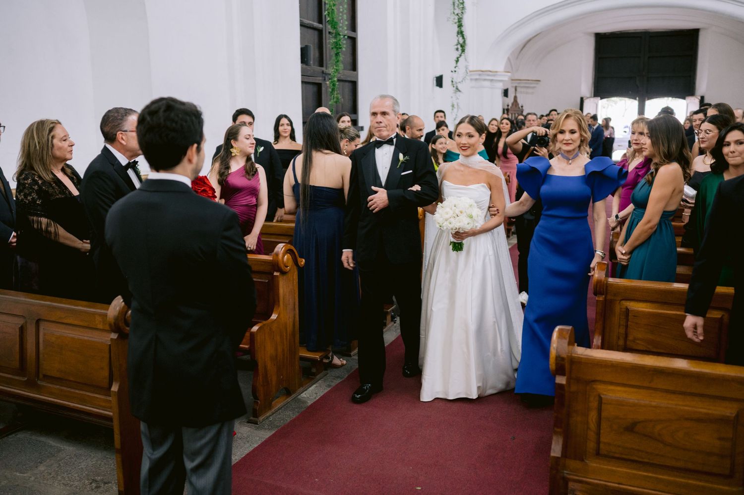 Wedding ceremony procession in church with guests in formal attire lining wooden pews.