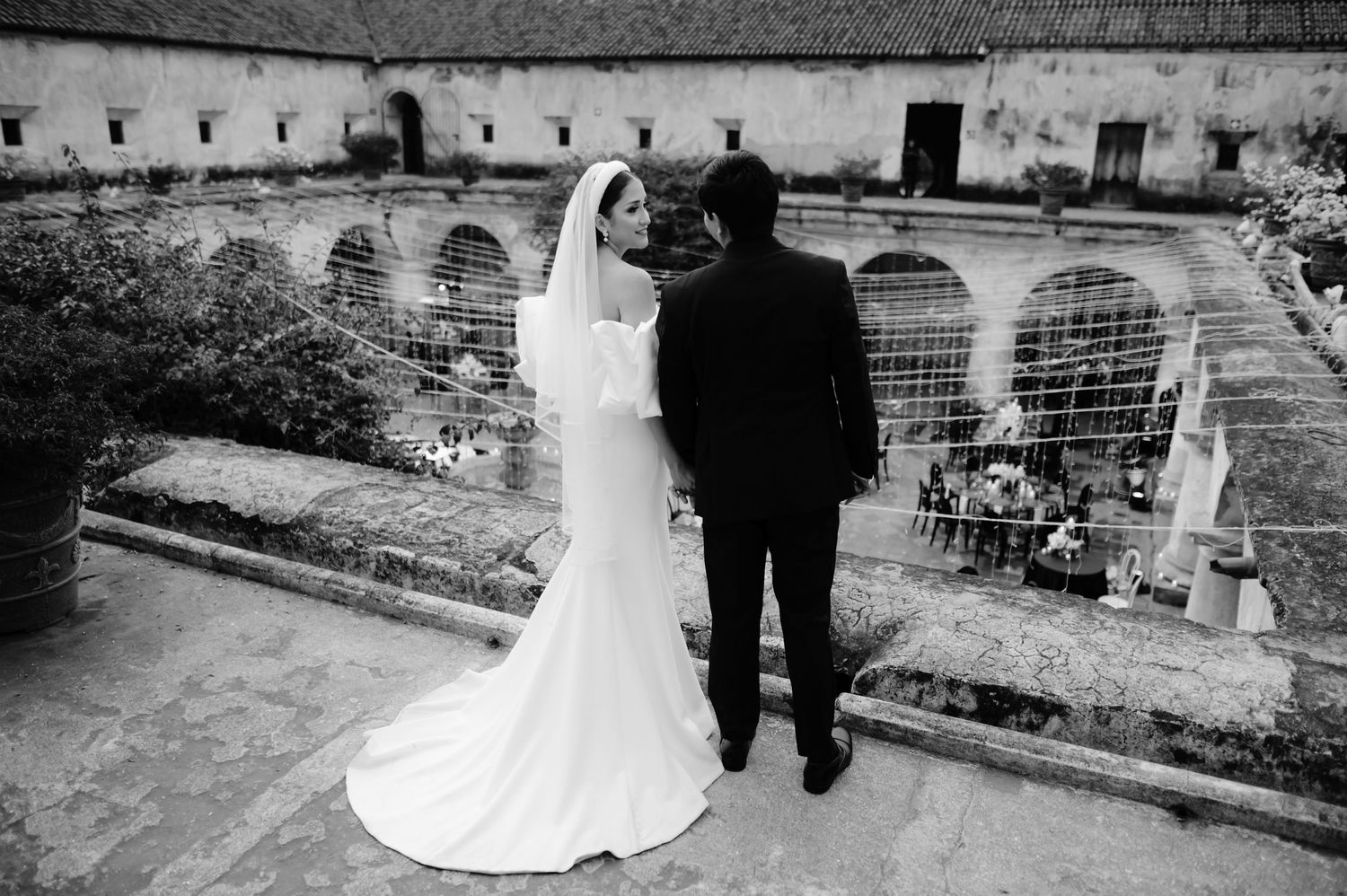 A wedding couple stands at a historic stone venue with arched architecture in black and white photography.