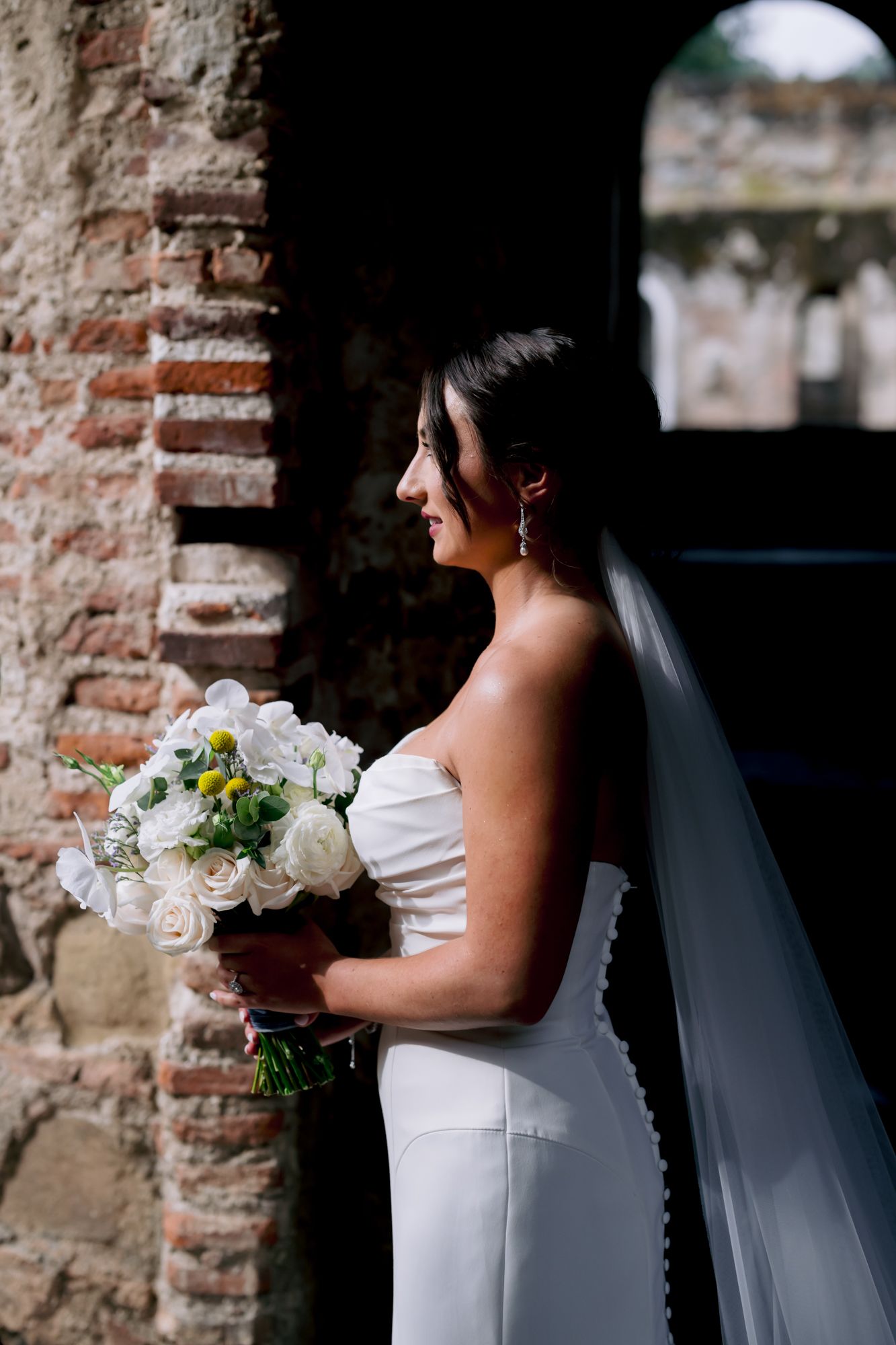 A bride in a white wedding dress holds a bouquet while standing against ancient brick ruins.