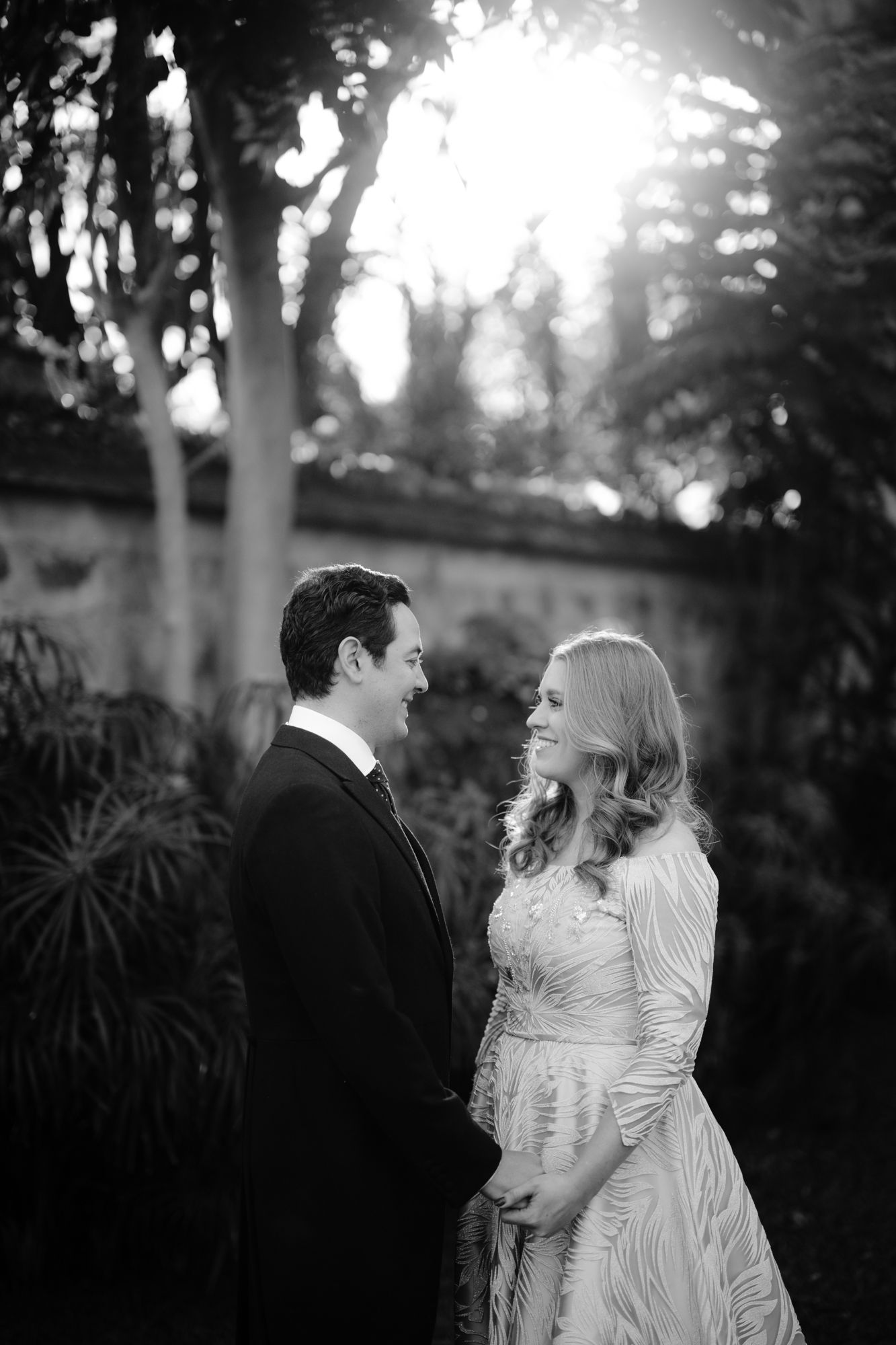 Black and white romantic portrait of couple in formal attire against garden background at sunset.