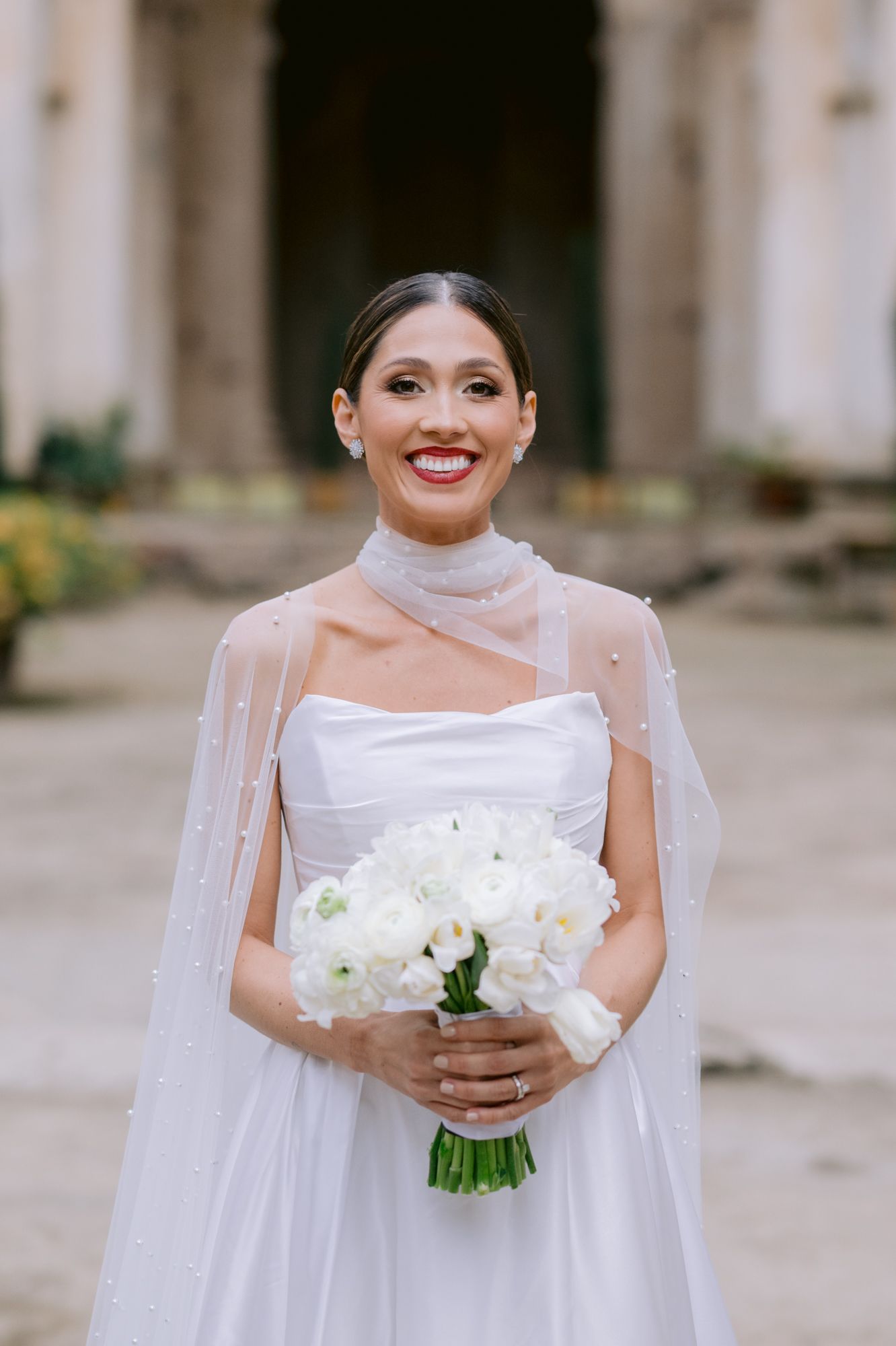 Bride in sheer cape dress holding white bouquet poses in church courtyard.