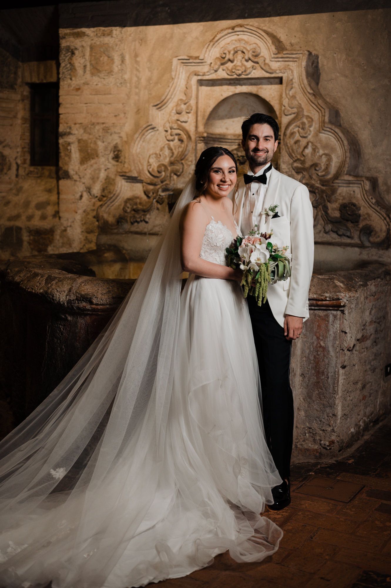 Elegant wedding portrait against ornate stone architecture with warm lighting.
