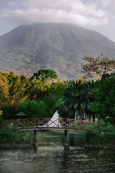 Scenic tropical landscape with wooden footbridge over water and misty volcano mountain in background near palm trees.