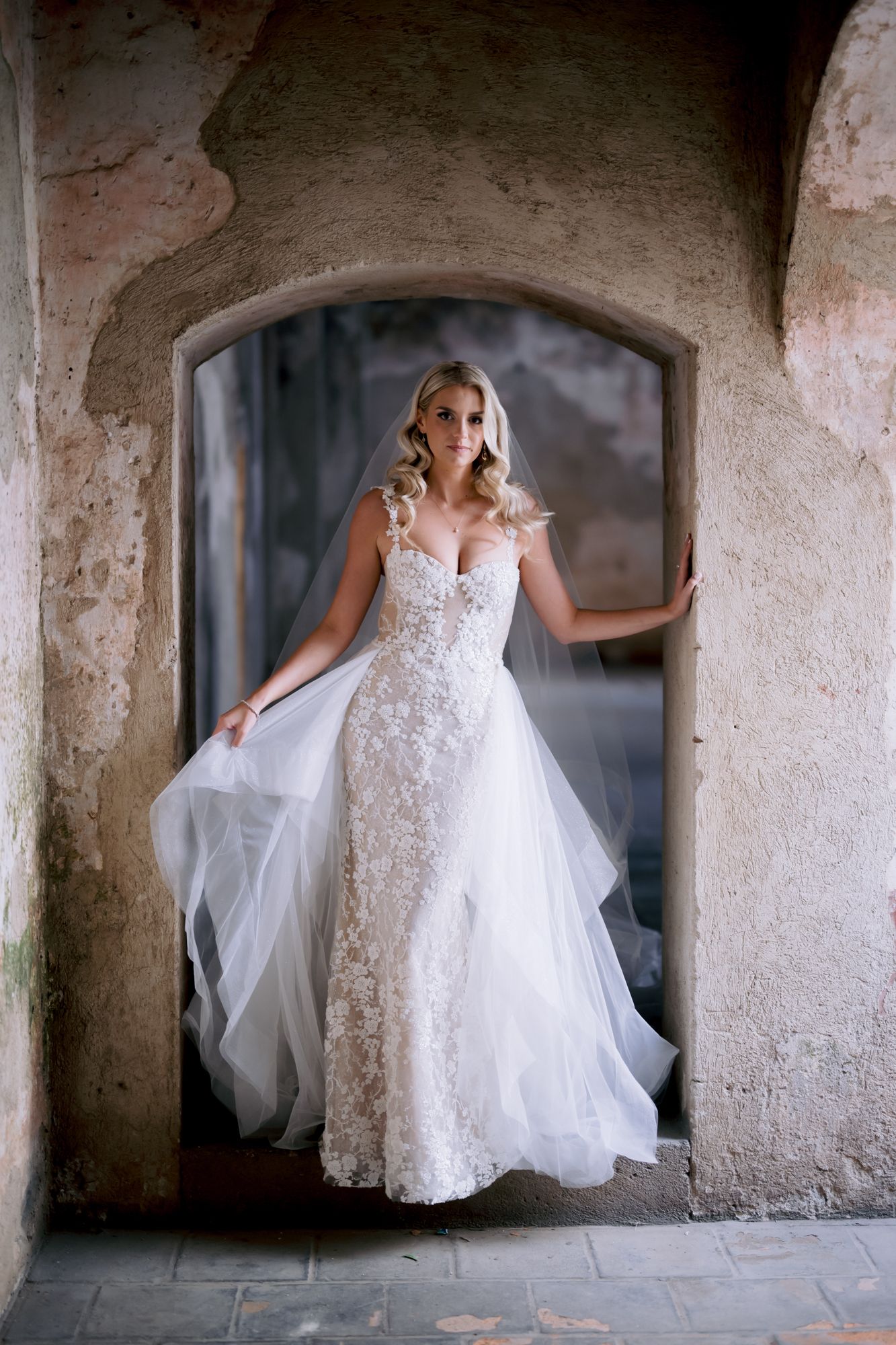 A bride in a detailed lace gown poses dramatically in a historic stone corridor.