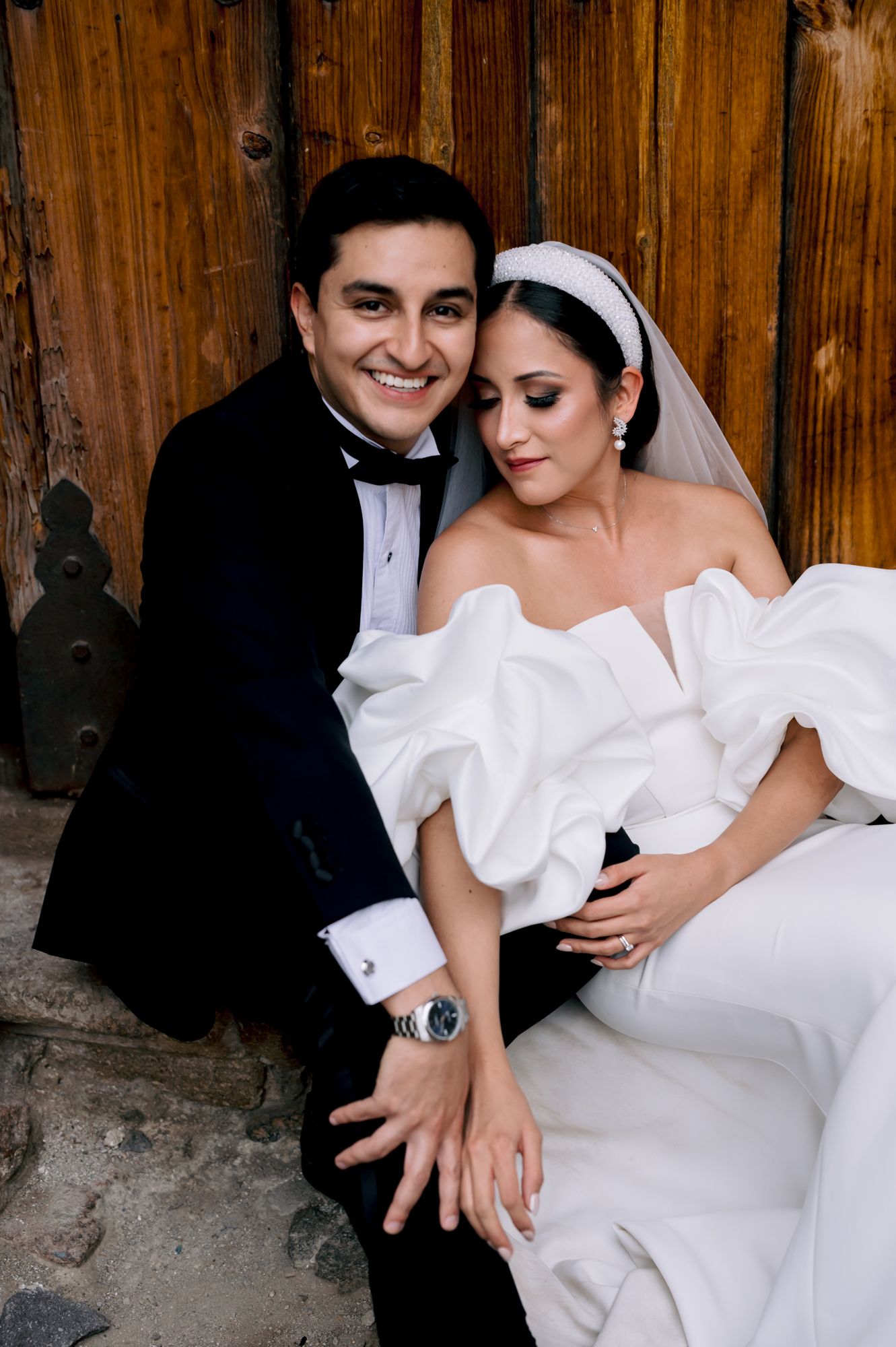 Wedding couple in classic black and white attire pose against rustic wooden door.