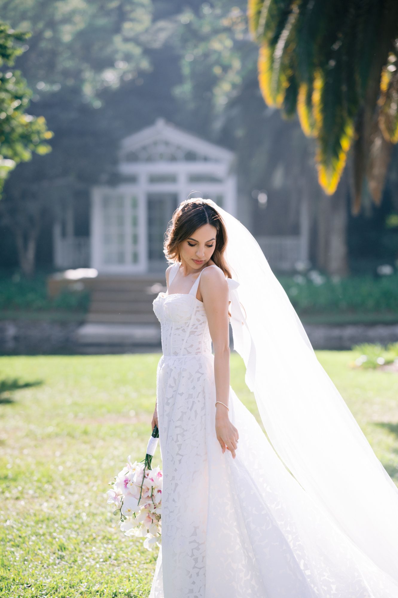 A bride in a detailed white gown walks through a garden with her flowing veil trailing behind.