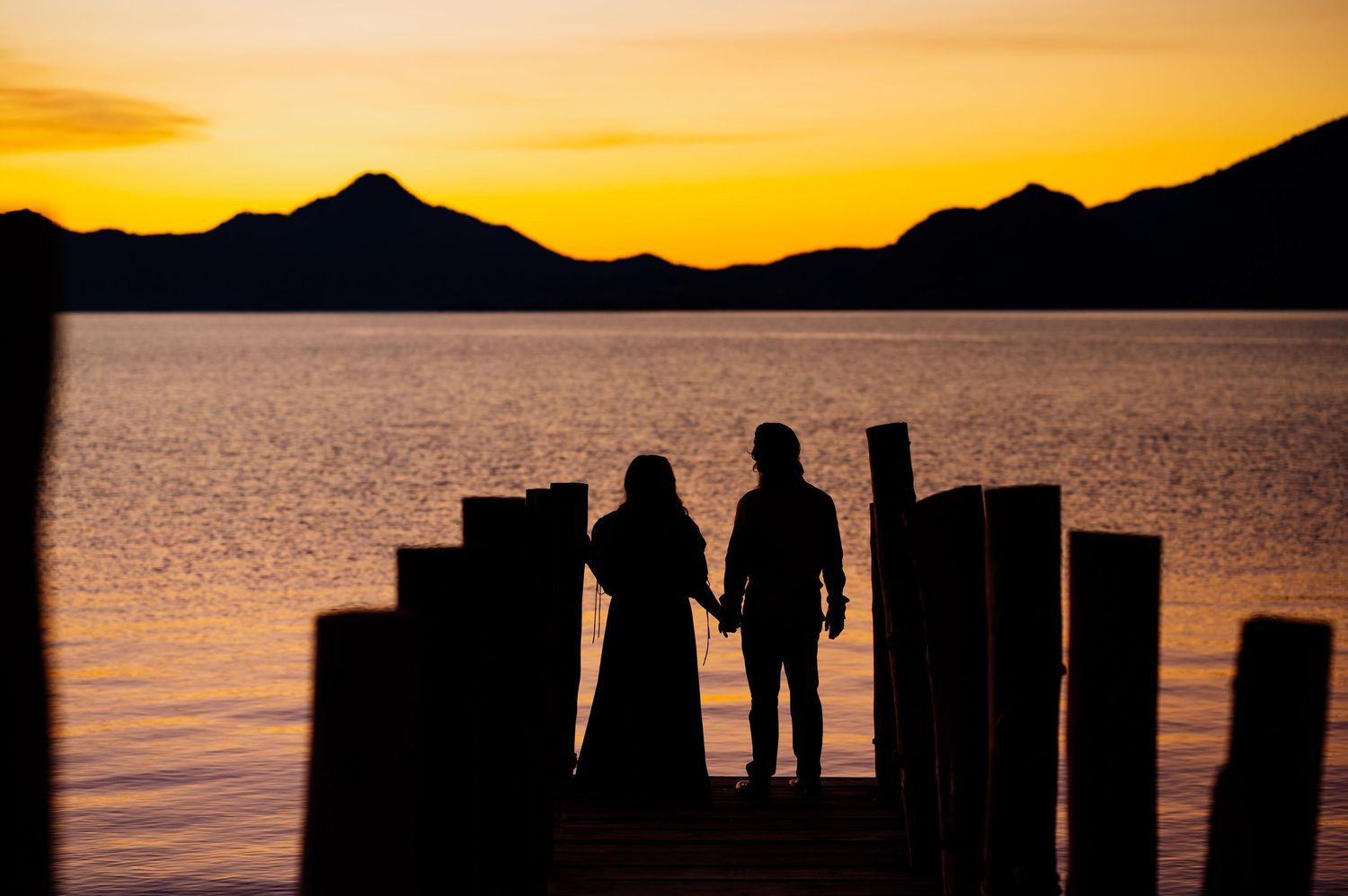 Silhouettes of people standing on a dock during golden sunset over mountain lake.