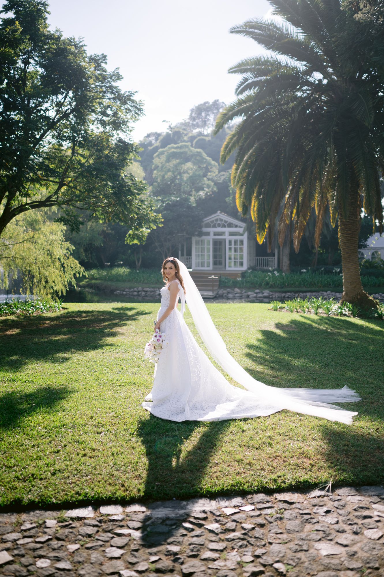 A flowing white wedding dress and veil twirl gracefully in a sunlit garden with palm trees and stone path.