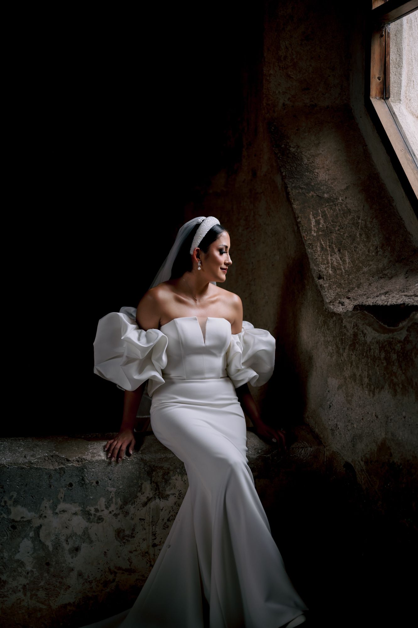 Bride in modern wedding dress with statement sleeves poses by window in dramatic lighting.