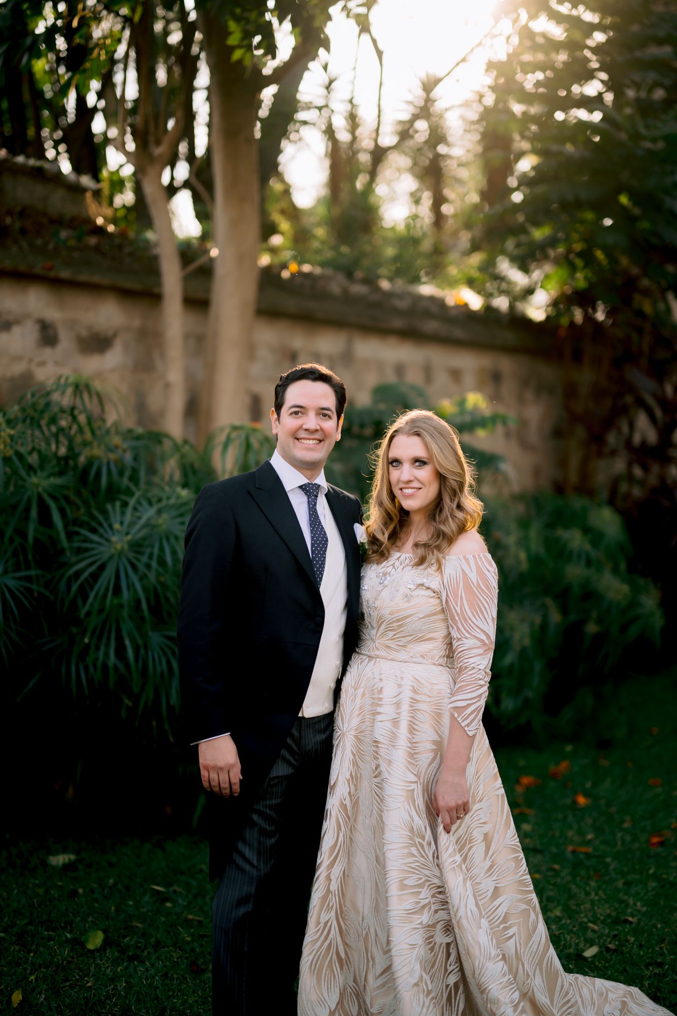 Bride and groom pose together in garden setting with stone wall and sunset lighting.