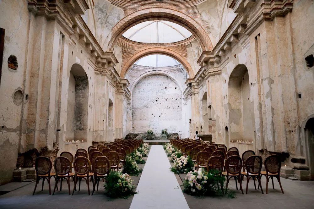 White aisle runner leads through rows of wooden chairs in an elegant old church with vaulted ceilings and arched windows.