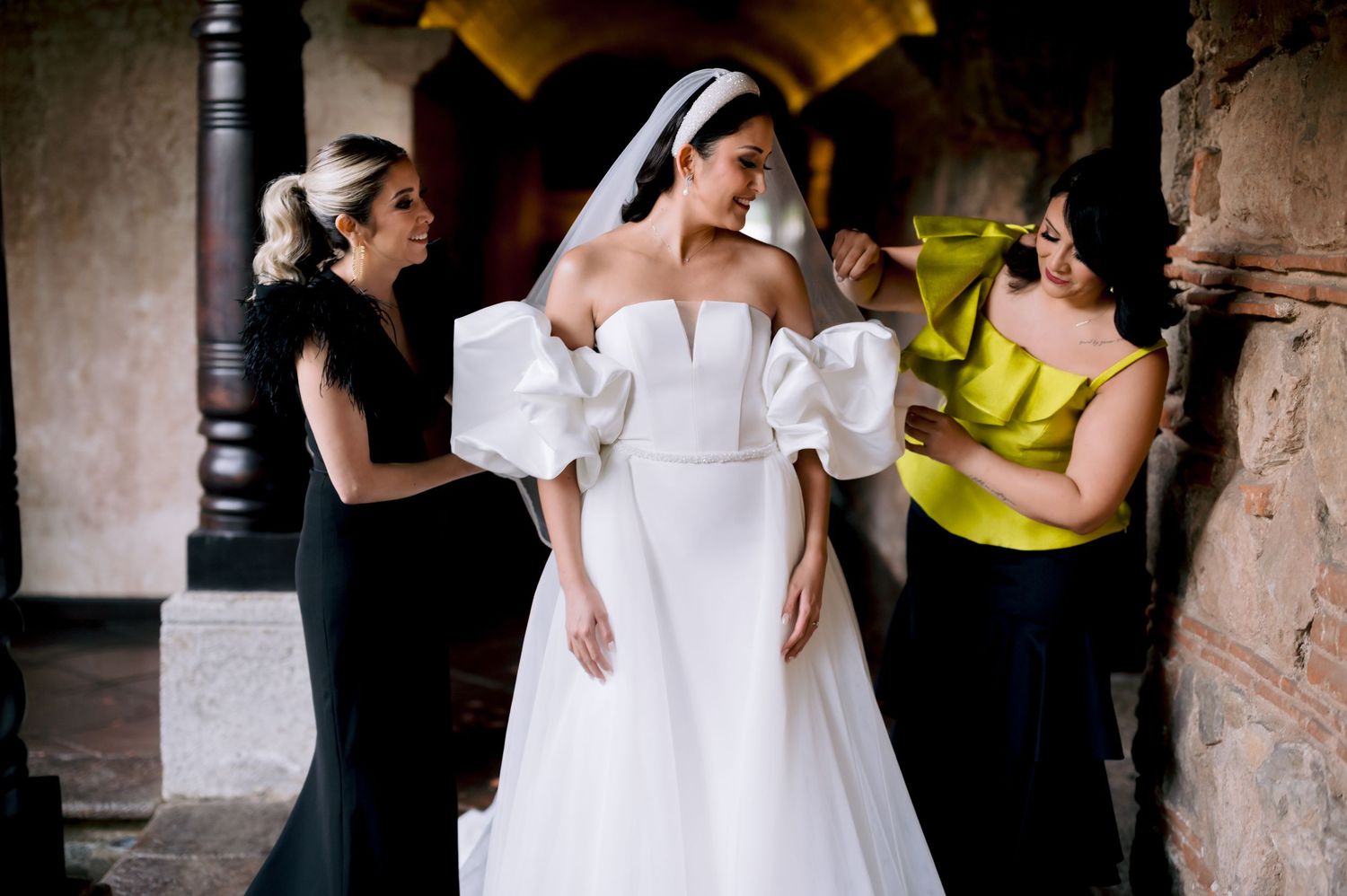Wedding dress fitting scene with assistants helping adjust the bride's gown in stone corridor.