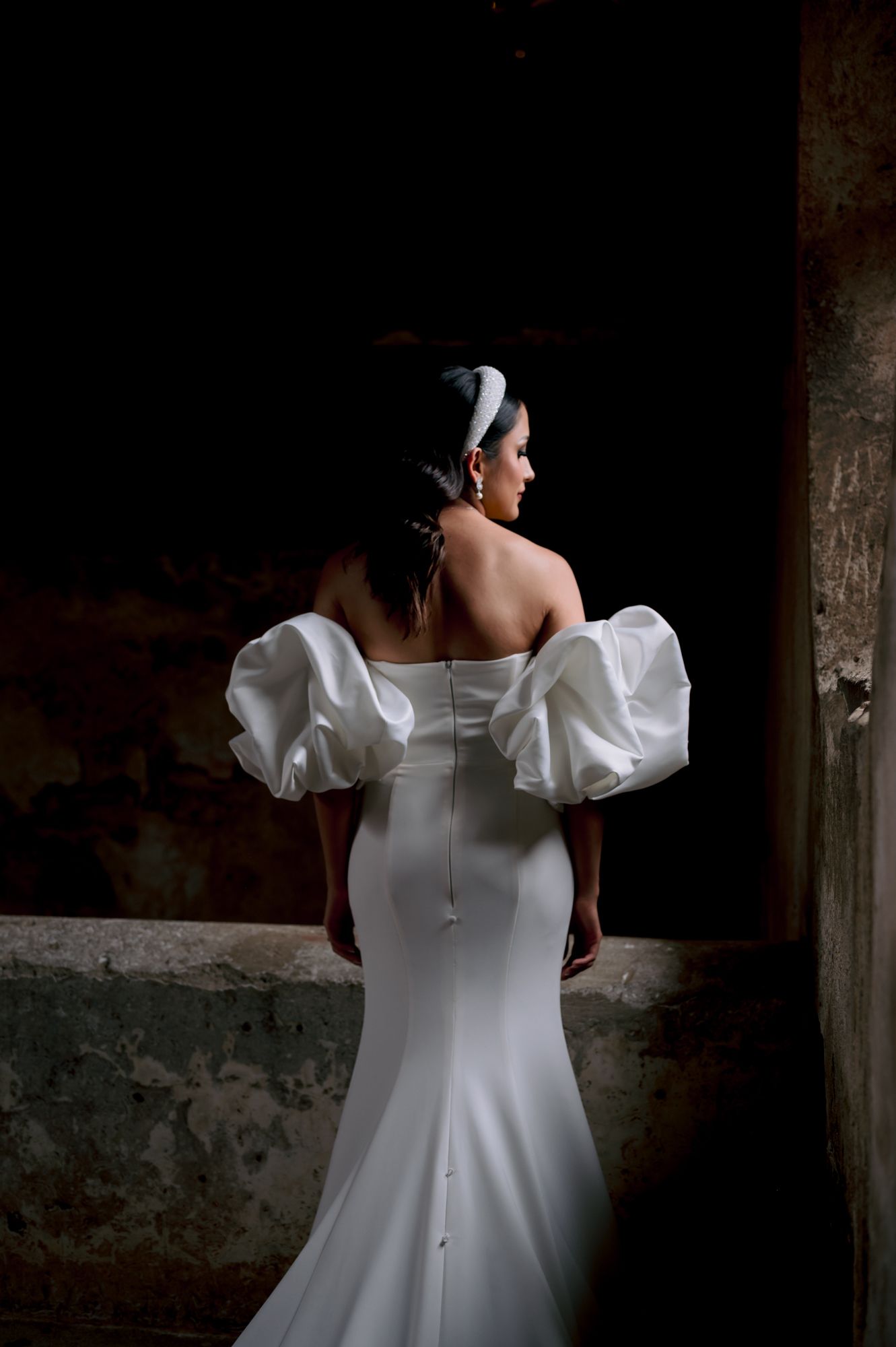 Back view of bride in fitted wedding gown with dramatic puff sleeves against dark background.