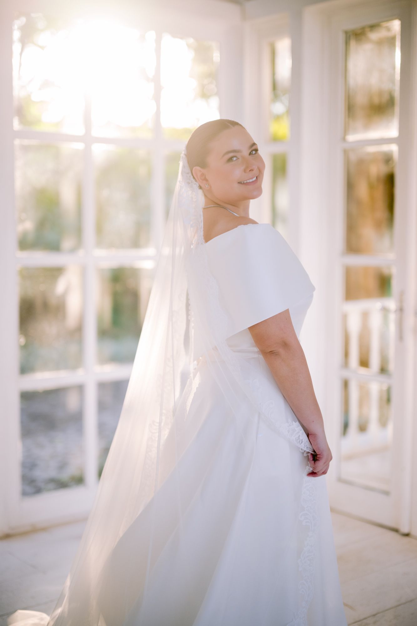 Radiant bride in off-shoulder wedding dress poses in sunlit white conservatory.