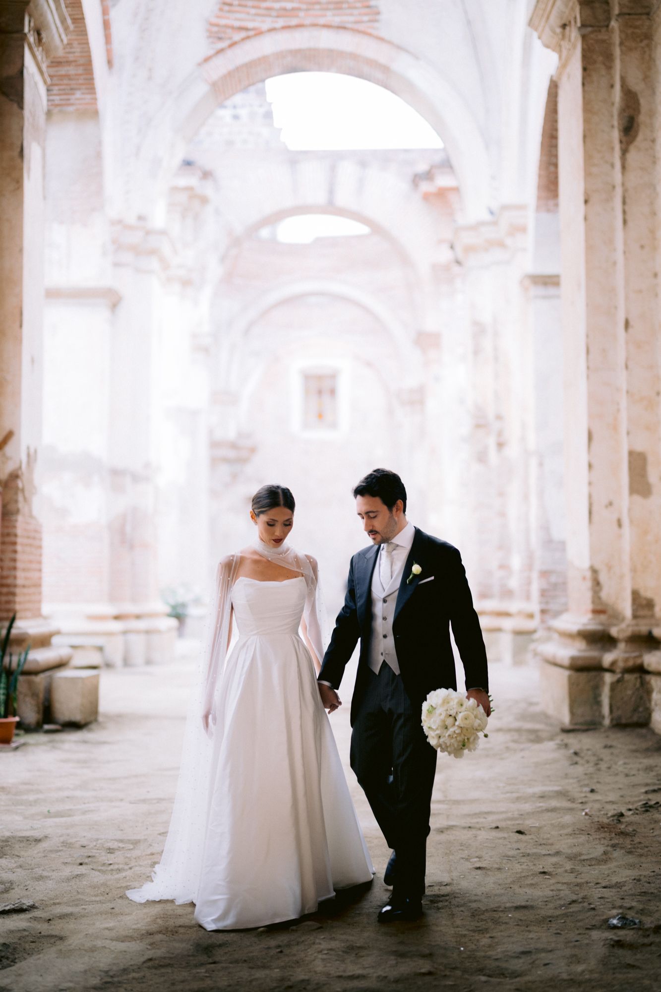 A bride and groom walking together in an elegant church with soaring arched columns and white bouquet.