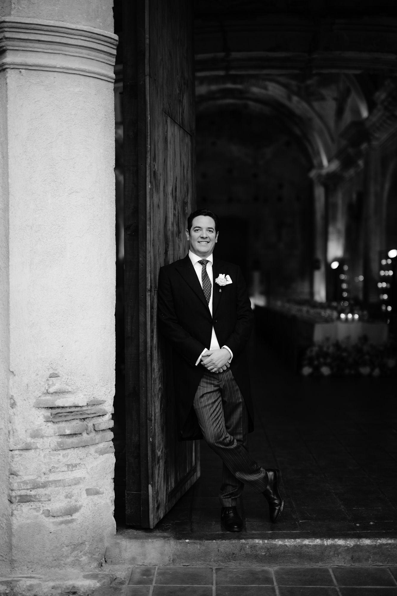 Groom in formal attire leans against stone column in dramatic architectural setting.