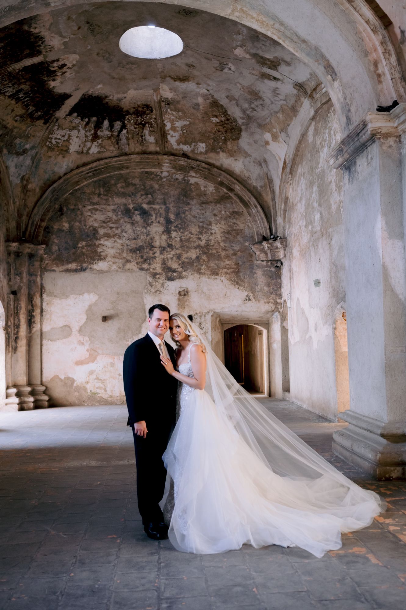 A wedding couple embraces in a rustic stone archway with dramatic lighting and flowing white gown.