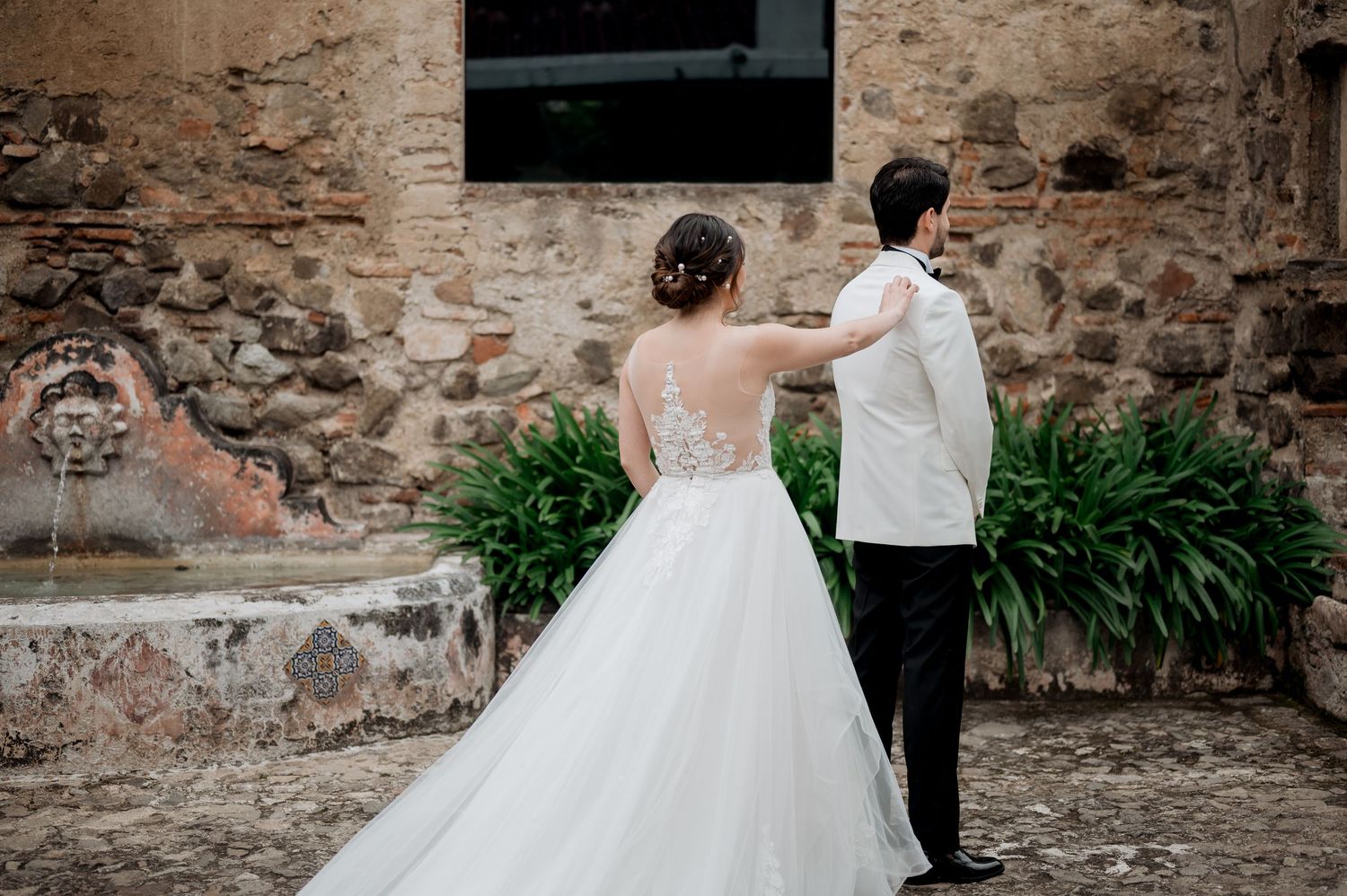 Couple shares intimate moment in white wedding attire against rustic stone wall backdrop.