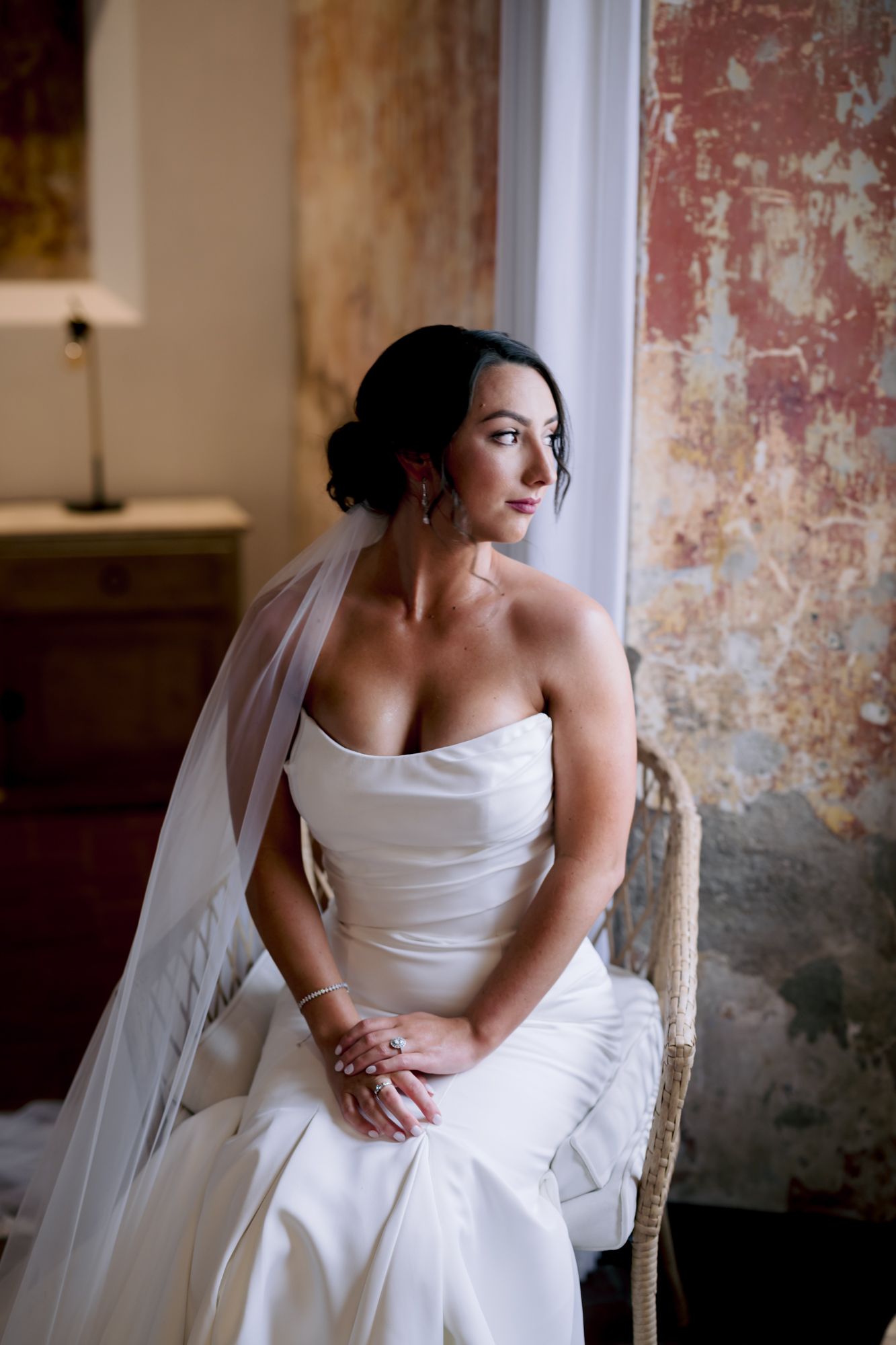 A bride in a strapless gown sits elegantly in front of a vintage wall.