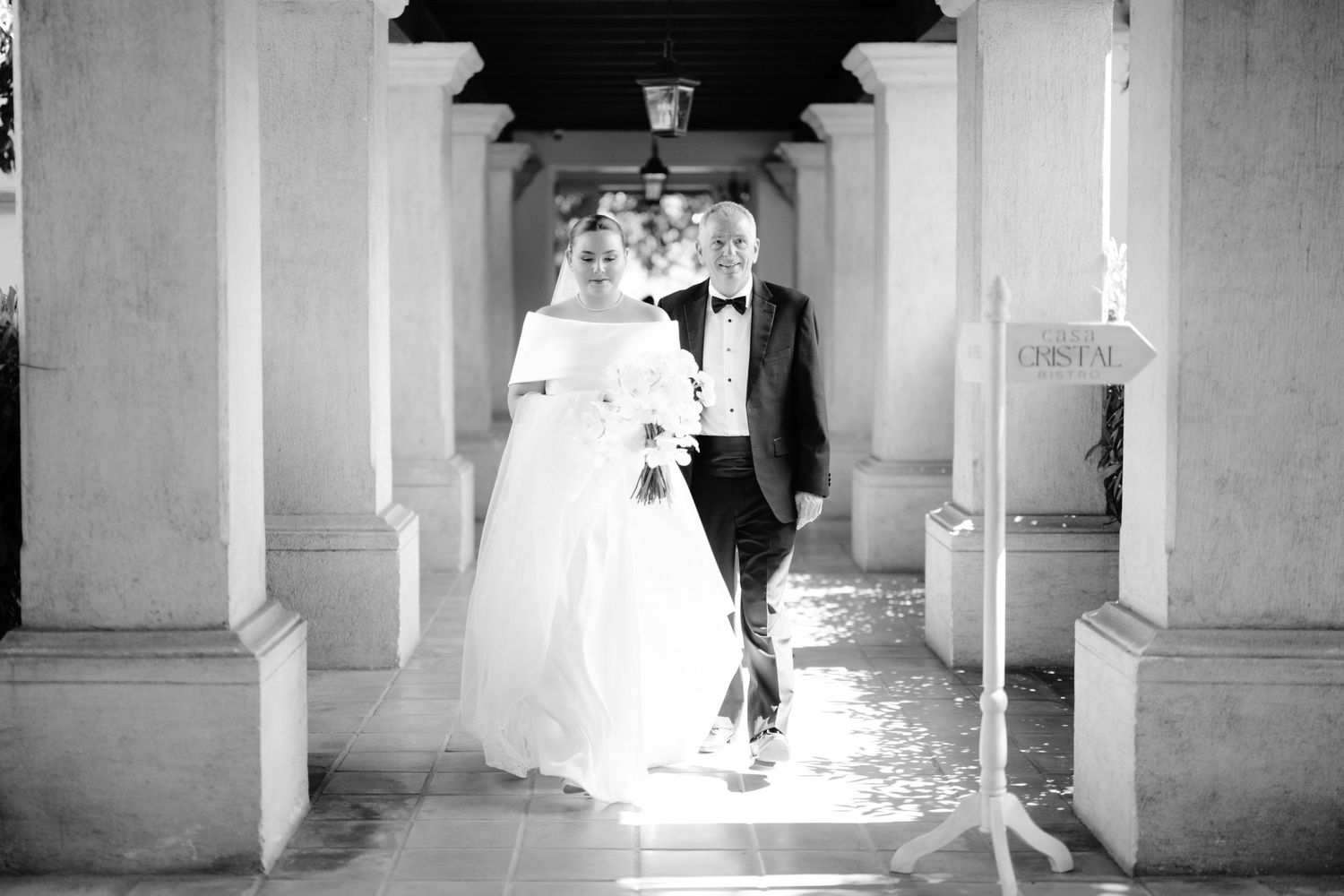 A black and white wedding photo capturing a walk through an elegant columned hallway.