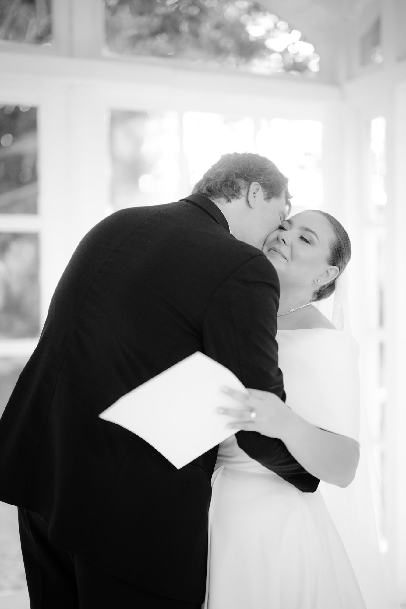 A romantic black and white wedding photo capturing a tender kiss between newlyweds in a sunlit room.
