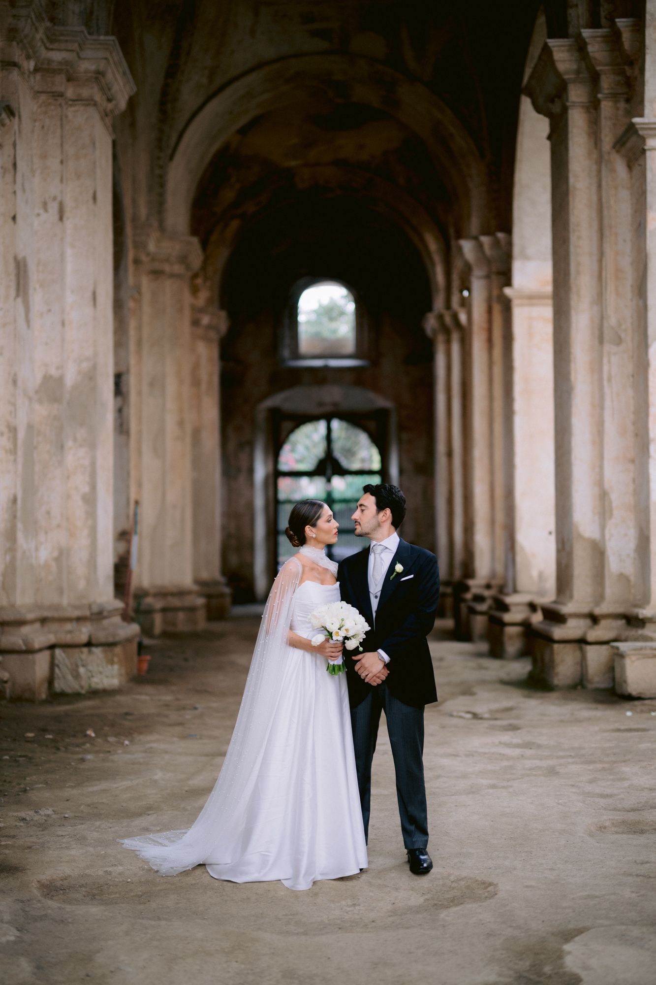 Wedding couple stands in elegant pose under historic stone arches holding a white bouquet.