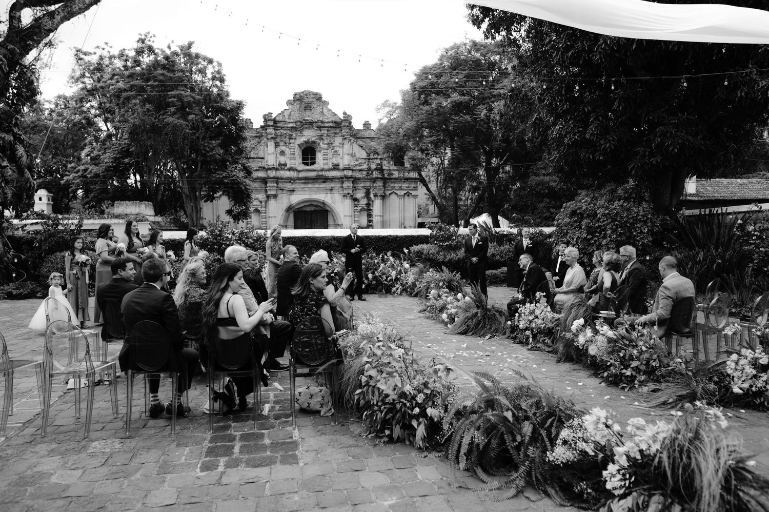 Black and white photo of wedding guests seated along garden path leading to historic church.
