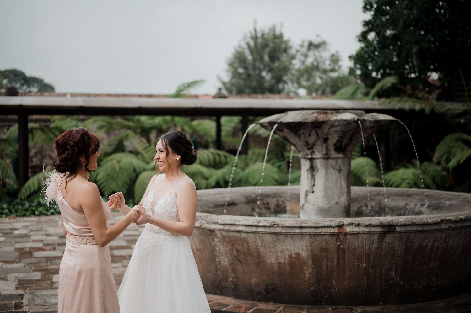 Two figures in blush and white dresses stand by an ornate stone fountain in a garden setting.