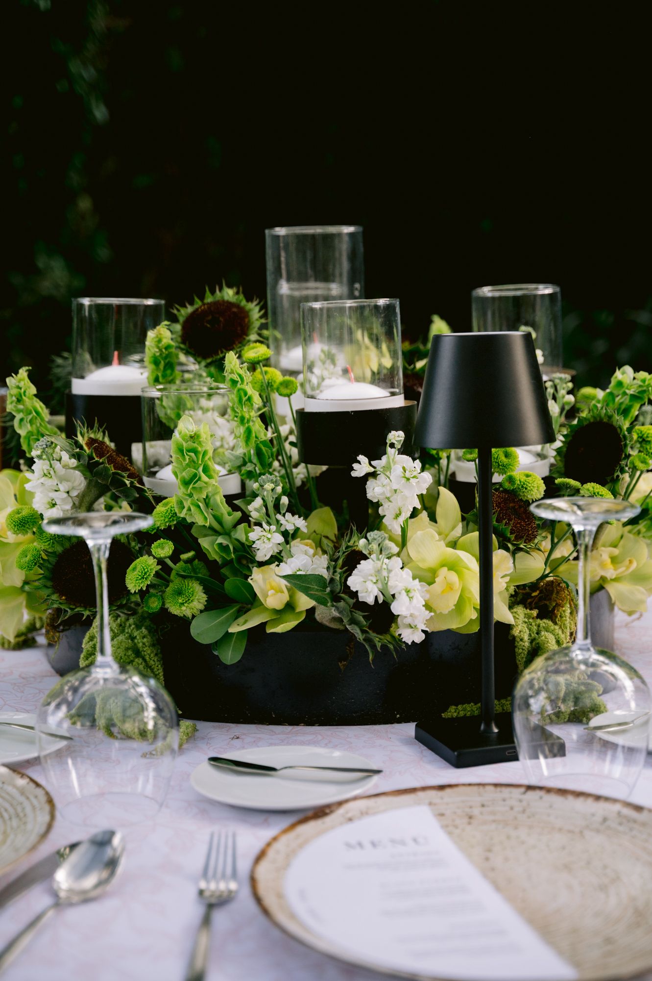 Elegant wedding table setting with black lampshades, white plates, and fresh green and white floral centerpieces.