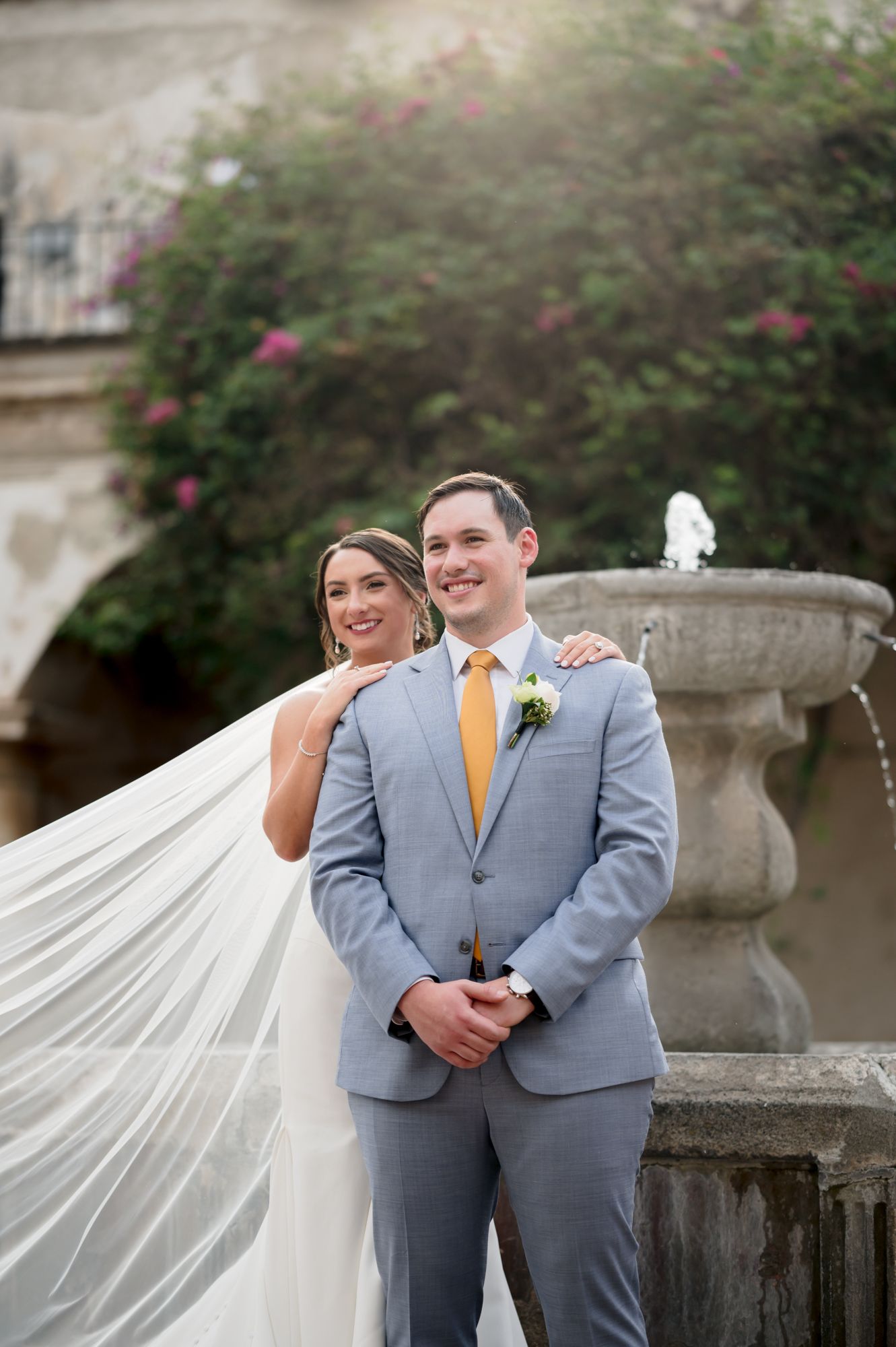 Elegant wedding portrait with flowing veil against garden fountain and floral backdrop.