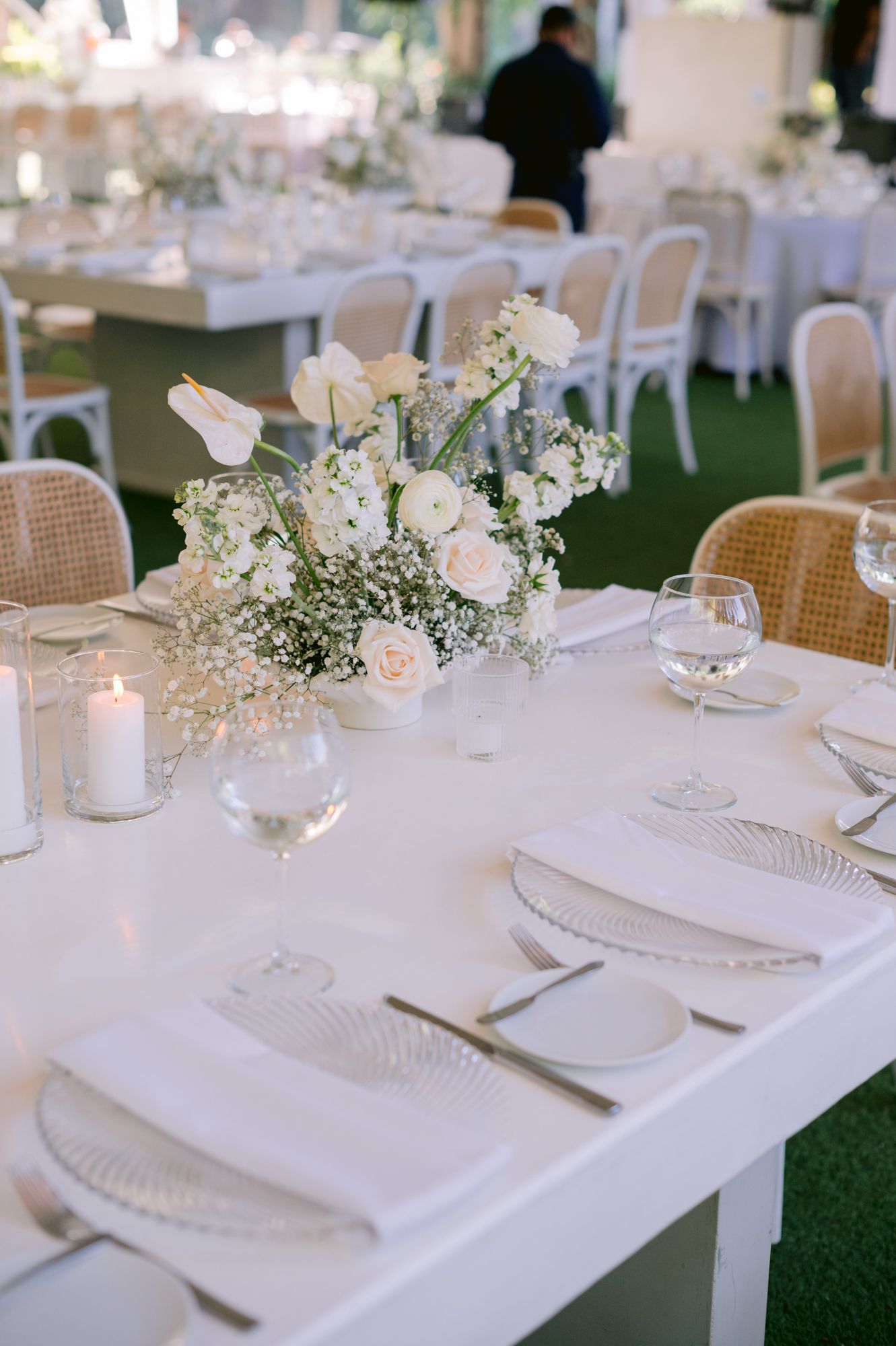 White wedding reception table with floral centerpieces, candles, and place settings on artificial grass.