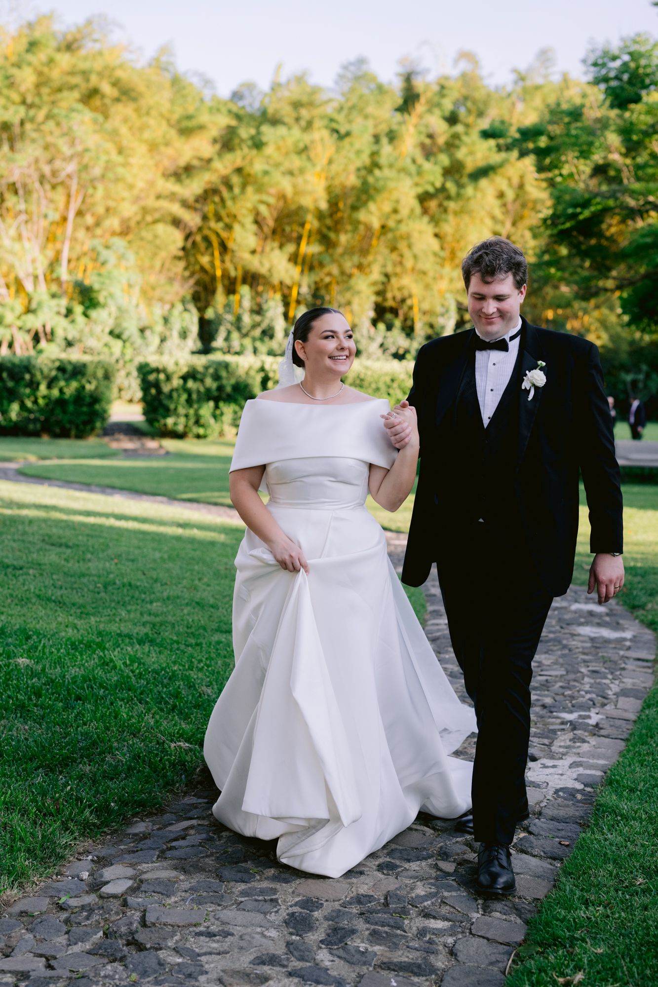 Wedding couple strolls together on a stone pathway through a sunny garden setting.