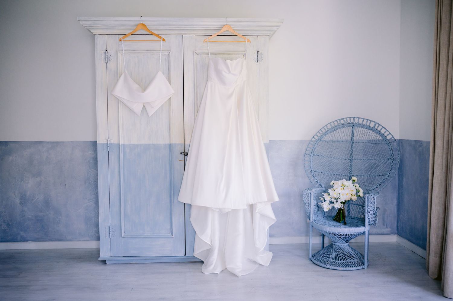 Elegant white wedding dress and attire hanging on wardrobe doors with blue walls and decorative chair.