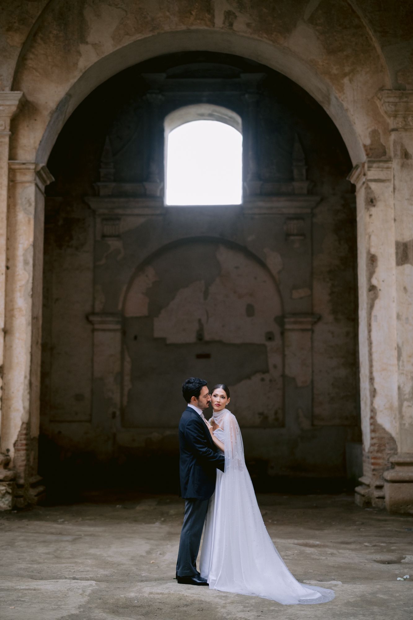 Wedding couple standing under stone archway in romantic architectural setting.