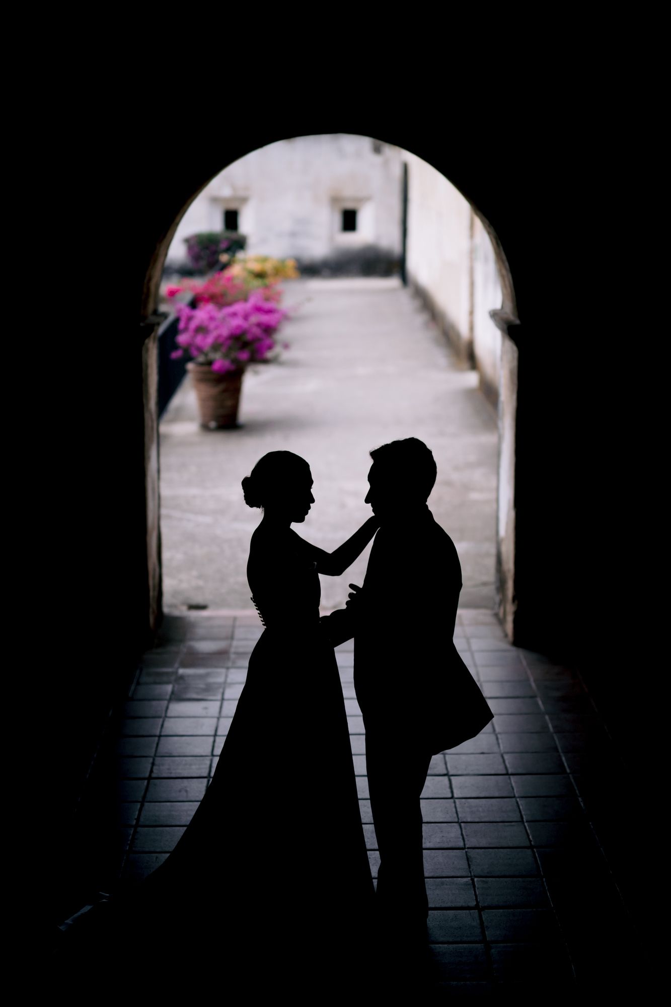 Silhouette of a couple in formal attire framed by an archway with pink flowers in the background.