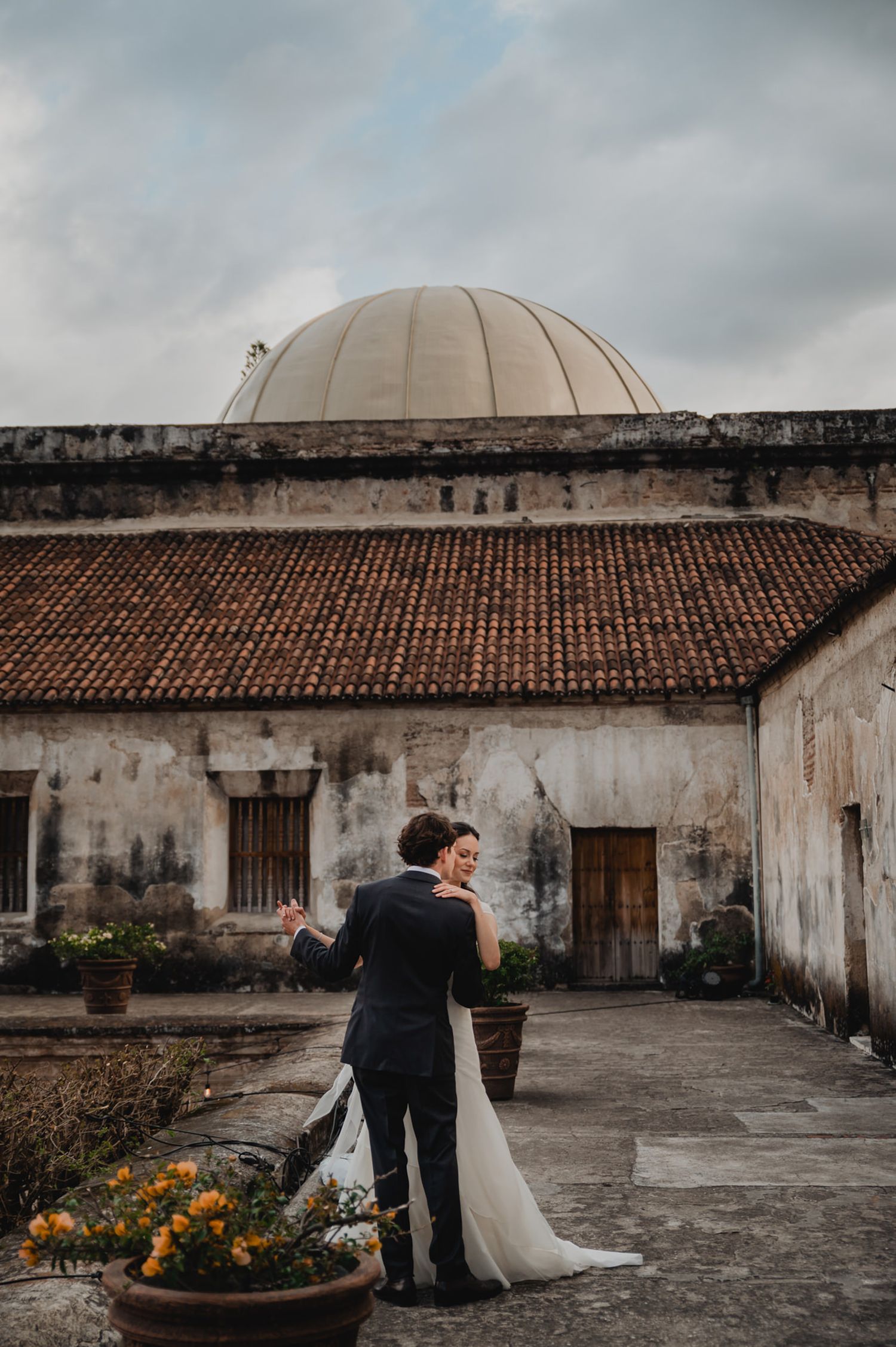 Couple embraces under domed architecture with weathered walls and flowering plants.