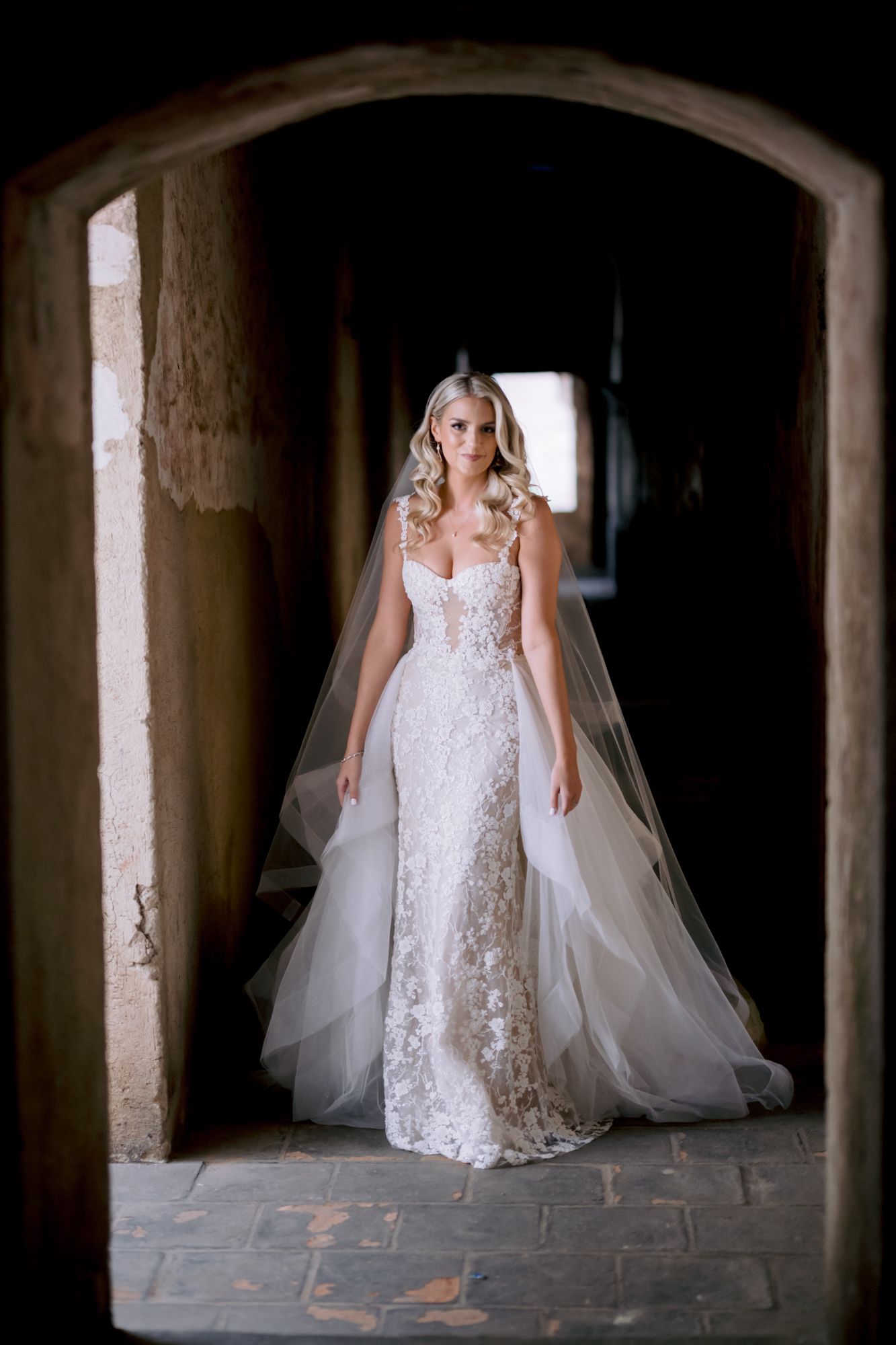 Bride wearing fitted lace gown and cathedral veil poses in historic stone corridor.