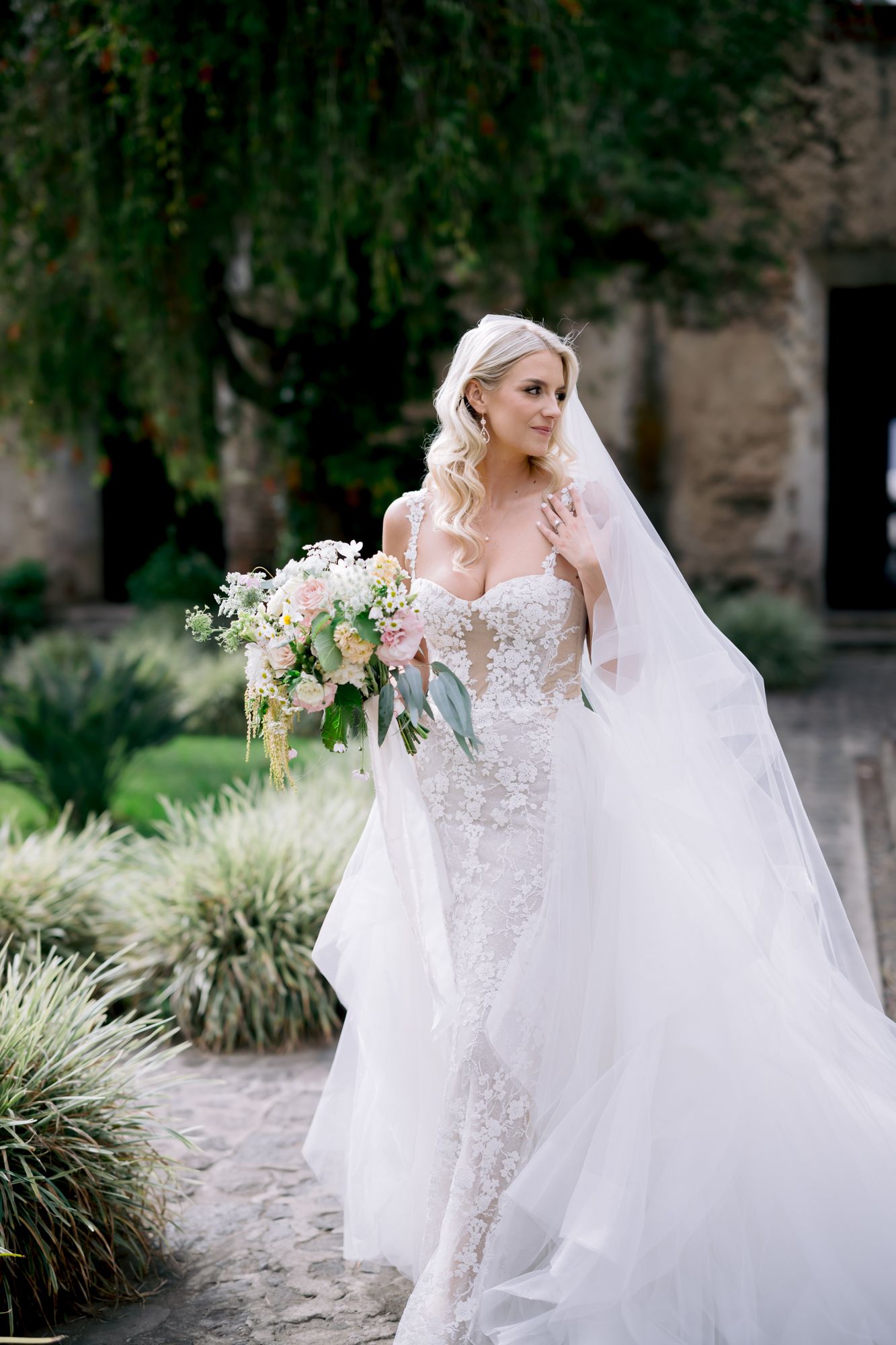 Bride in fitted lace gown holds bouquet while standing in garden setting.
