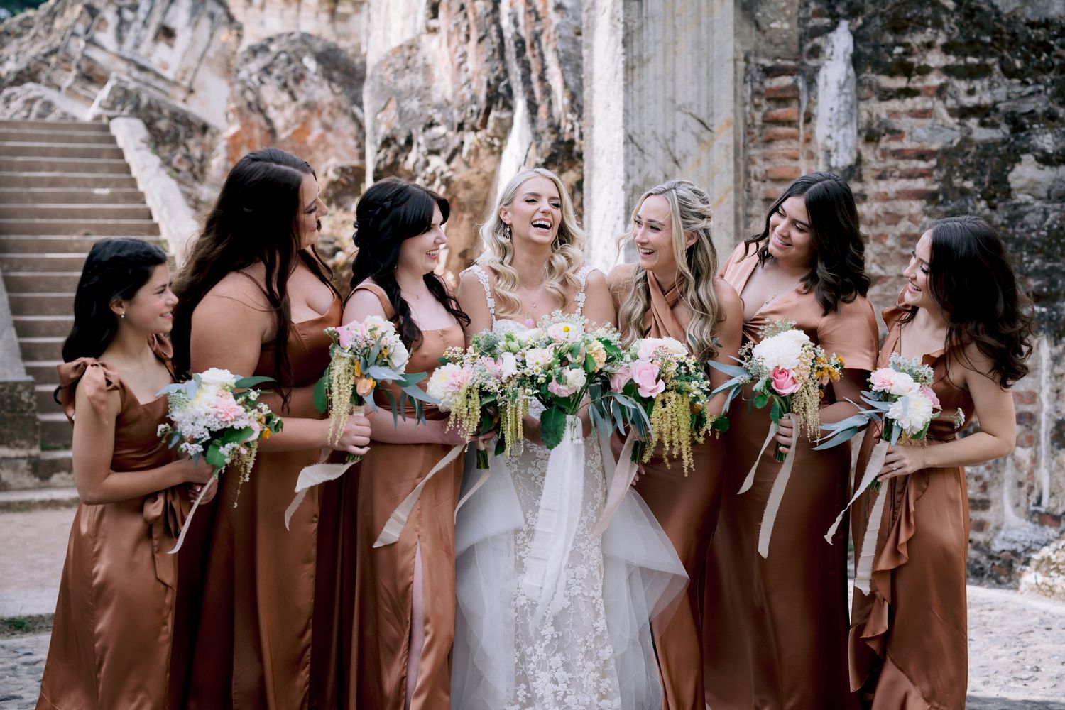 Bridesmaids in copper gowns surround bride for photos at historic venue.