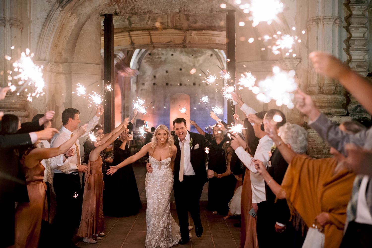 Newlyweds celebrate with guests holding sparklers during their grand exit at an elegant indoor venue.