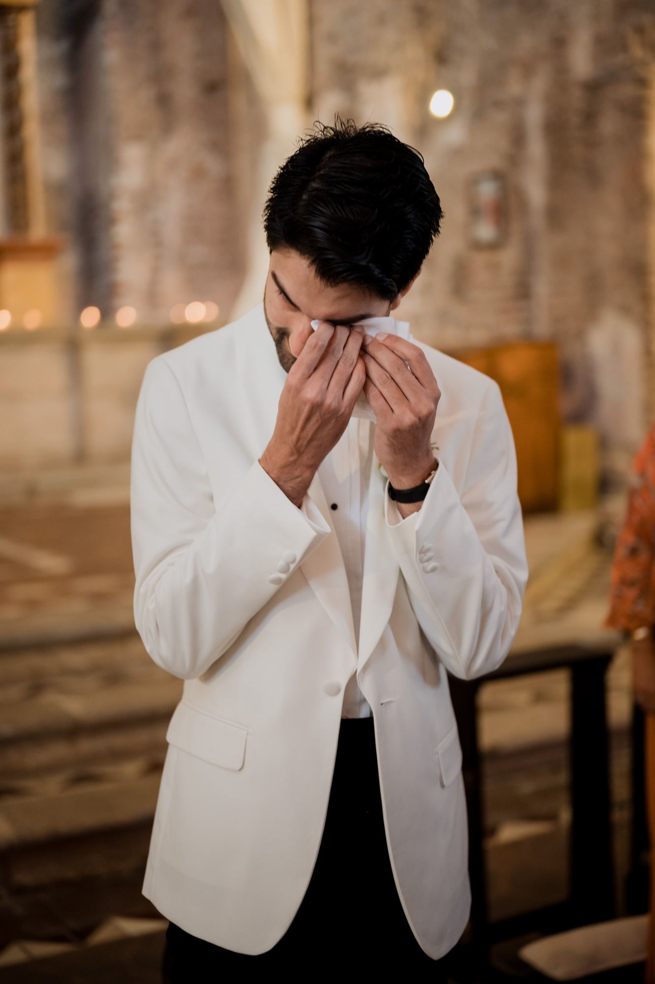 An emotional moment captured as a groom in white jacket covers his face with his hands.