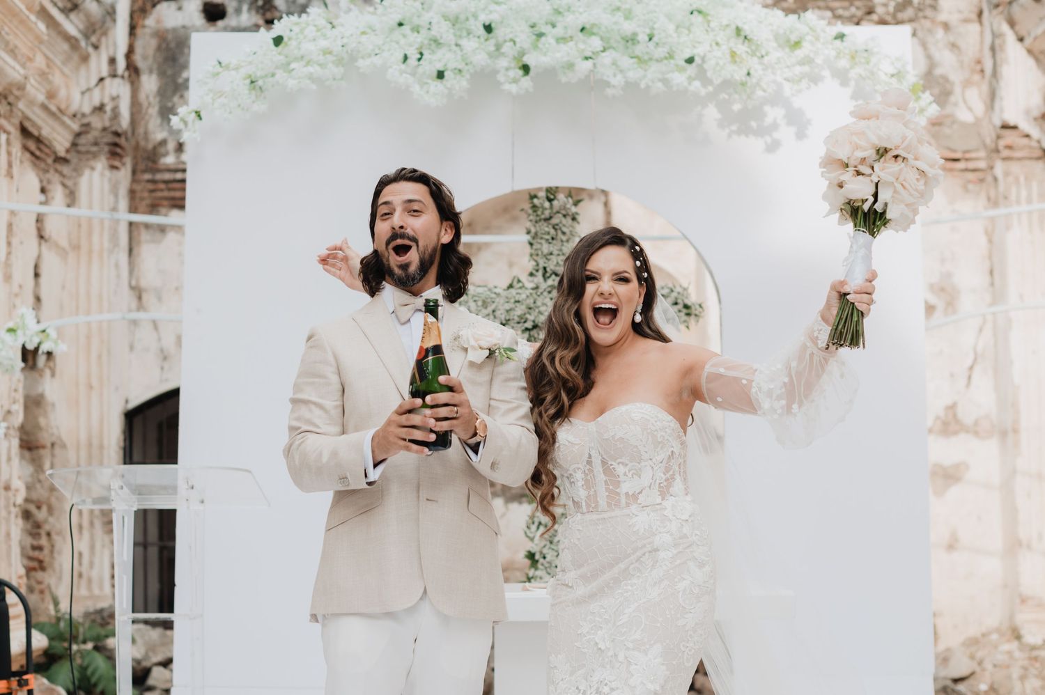 Joyful wedding party celebrates under white stone archways with confetti flying through the air.